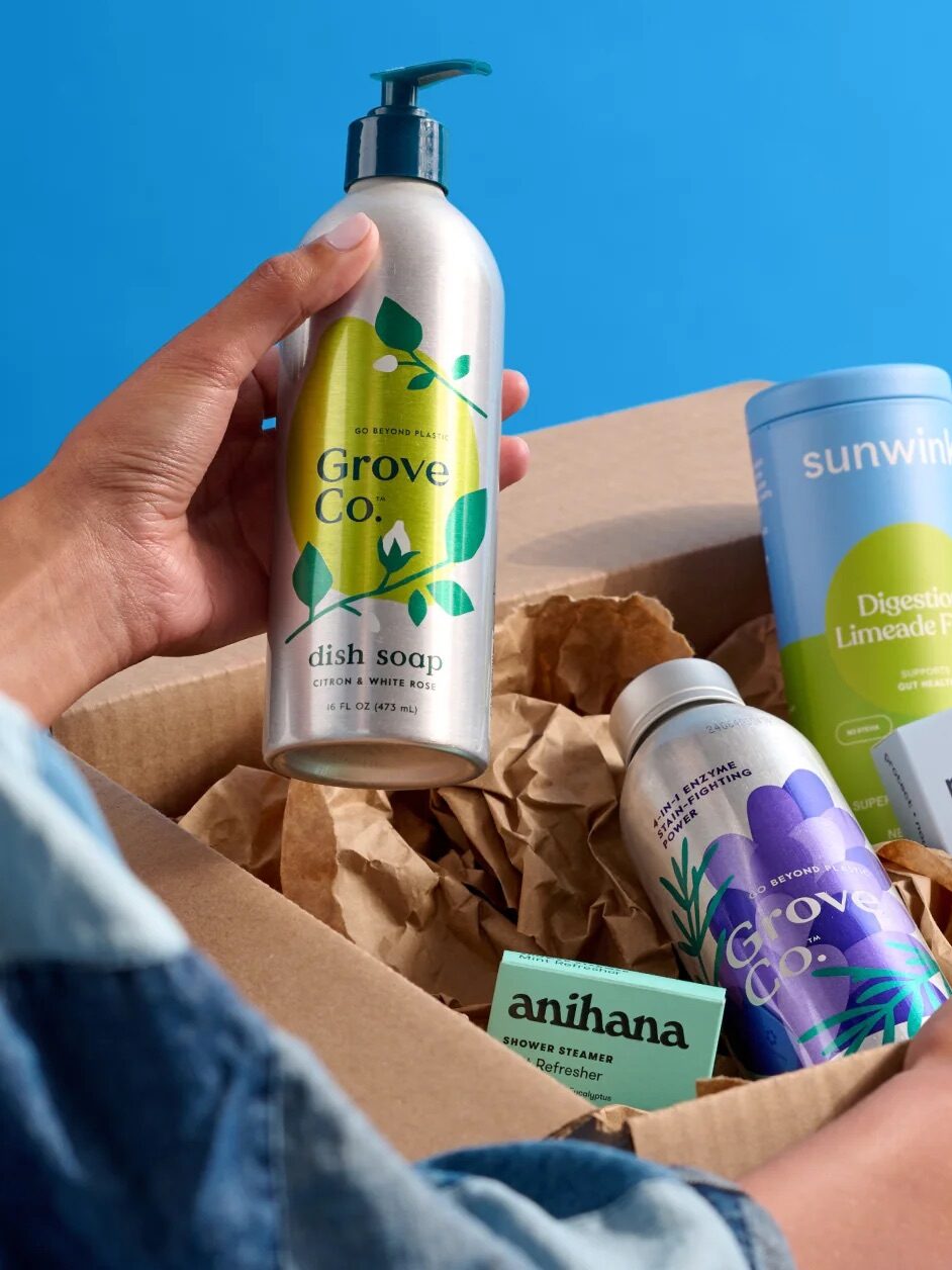 A person opens a cardboard box from online shopping, revealing assorted eco-friendly products like hand soap, dog waste bags, deodorant, paw wipes, and a small green box against a blue background with the Grove logo.