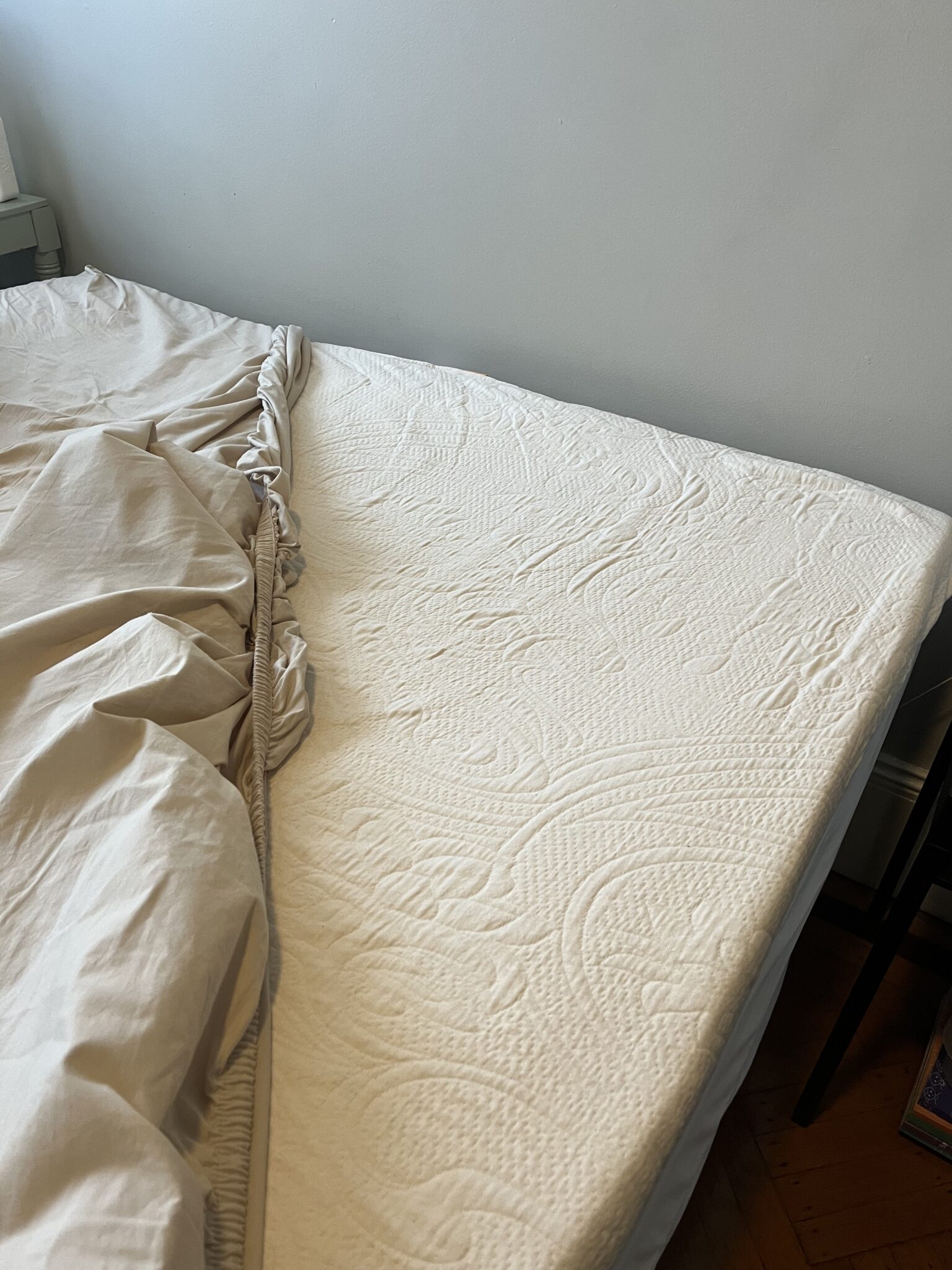 A bed with the fitted sheet partially pulled off, exposing a white mattress with a textured pattern next to a beige sheet.