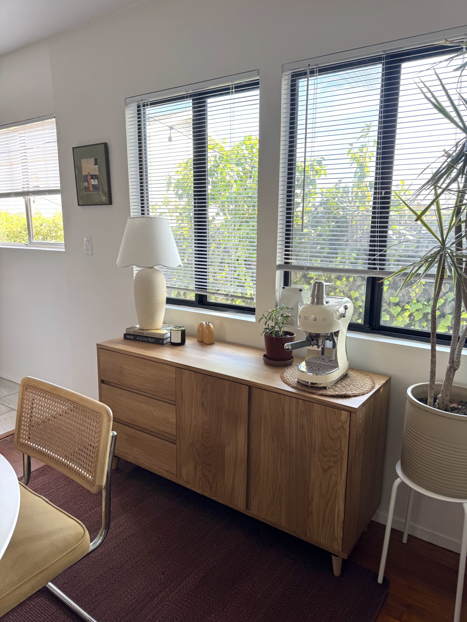 A wooden sideboard holds a lamp, books, potted plants, and a stand mixer. A chair and tall plant are nearby; windows with blinds let in natural light.