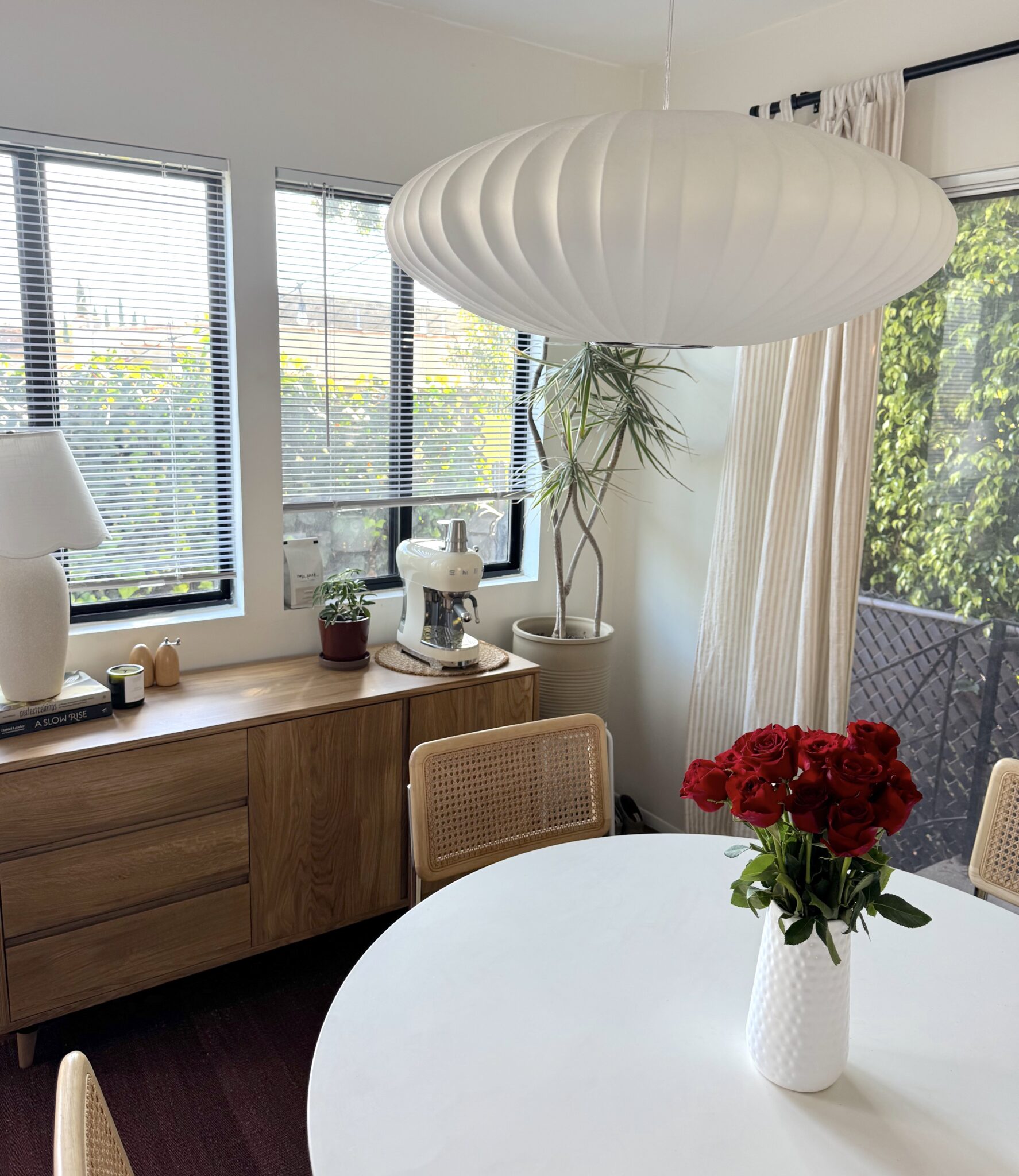 A white round dining table with a vase of red roses is surrounded by wooden chairs; a sideboard, coffee machine, and potted plant are near windows with blinds and outdoor greenery visible.