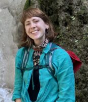 A person wearing a teal shirt, pink shorts, and a backpack stands in front of a rocky waterfall, smiling at the camera.