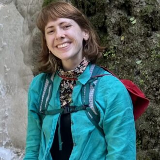 A person wearing a teal shirt, pink shorts, and a backpack stands in front of a rocky waterfall, smiling at the camera.