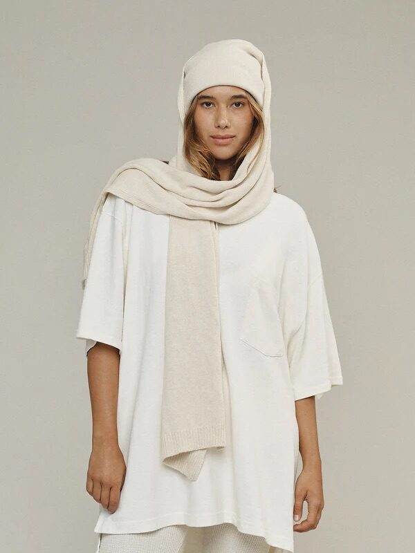 A woman stands against a plain background wearing an oversized white T-shirt, white knit pants, and a matching beige knit scarf and hat.