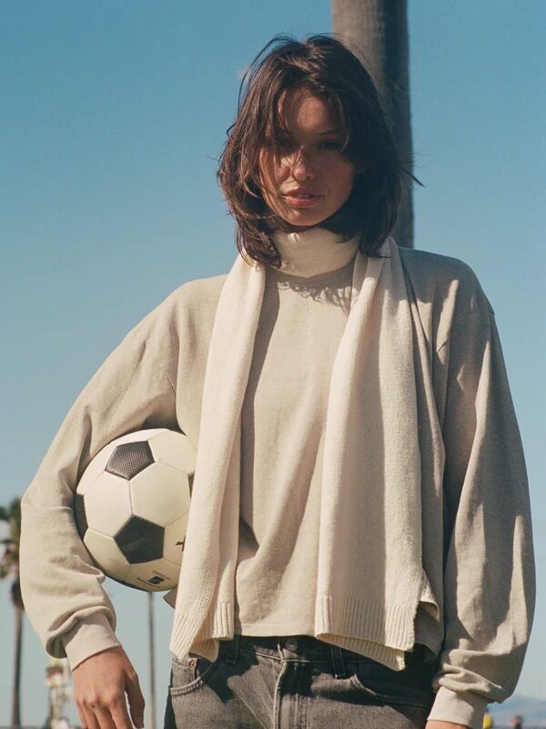Woman wearing a light sweater and scarf stands outdoors holding a soccer ball under her arm, with a clear sky and palm trees in the background.