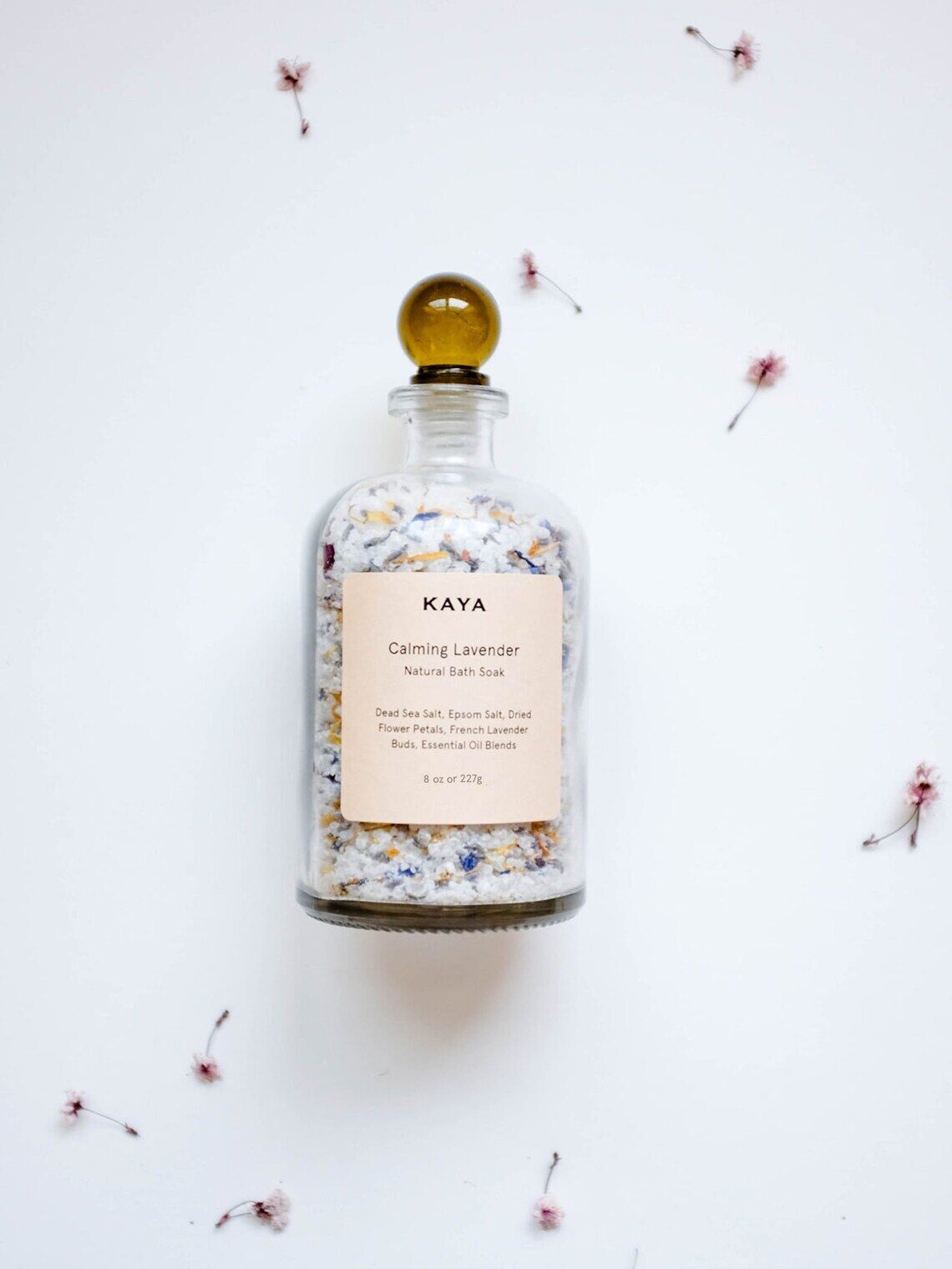 A glass bottle labeled "Kaya Calming Lavender Natural Bath Soak" is filled with bath salts and surrounded by small scattered dried flowers on a white background.