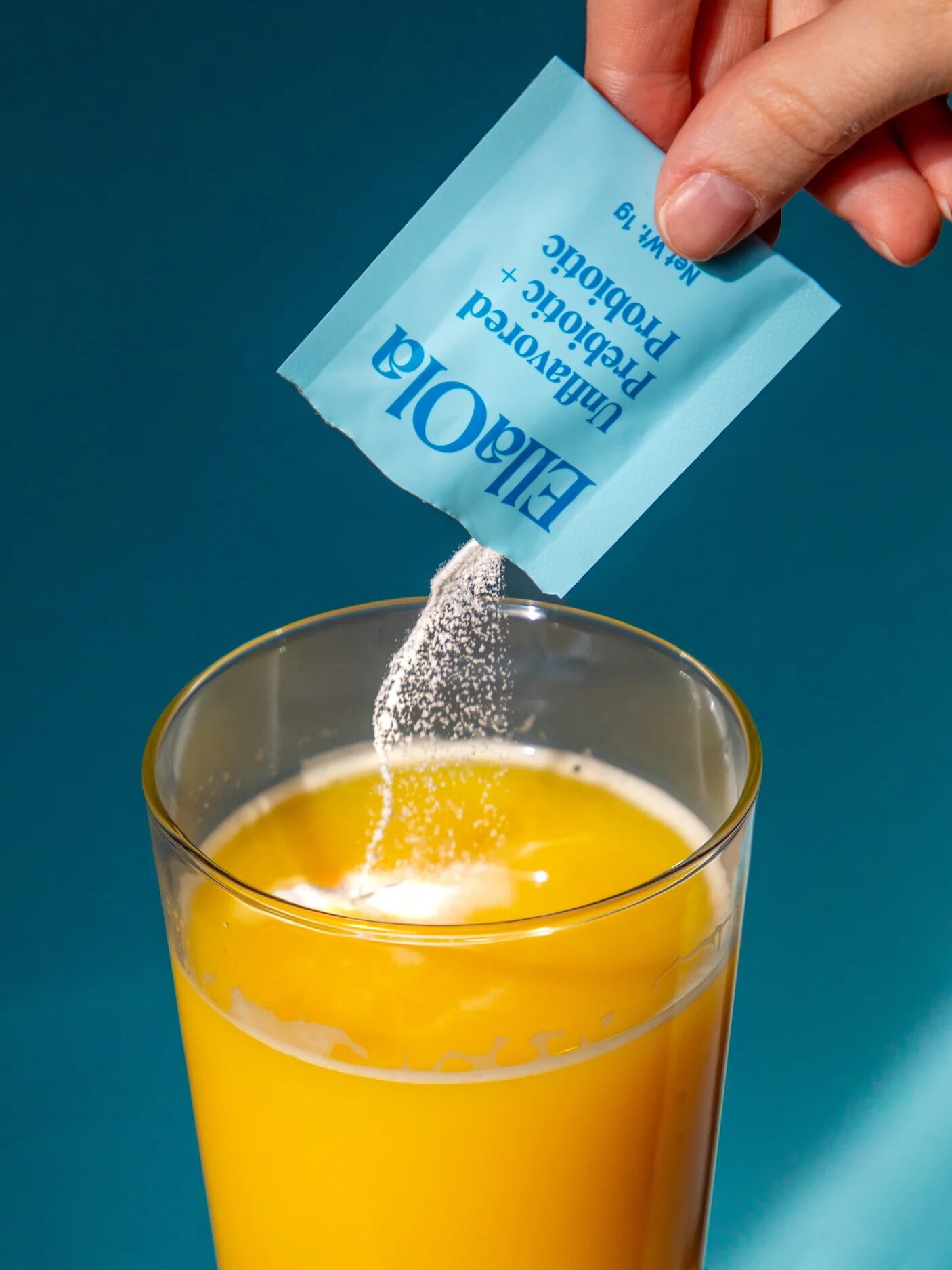 A hand pours powder from a blue packet labeled "Unflavored Prebiotic + Probiotic" into a glass of orange juice against a blue background.