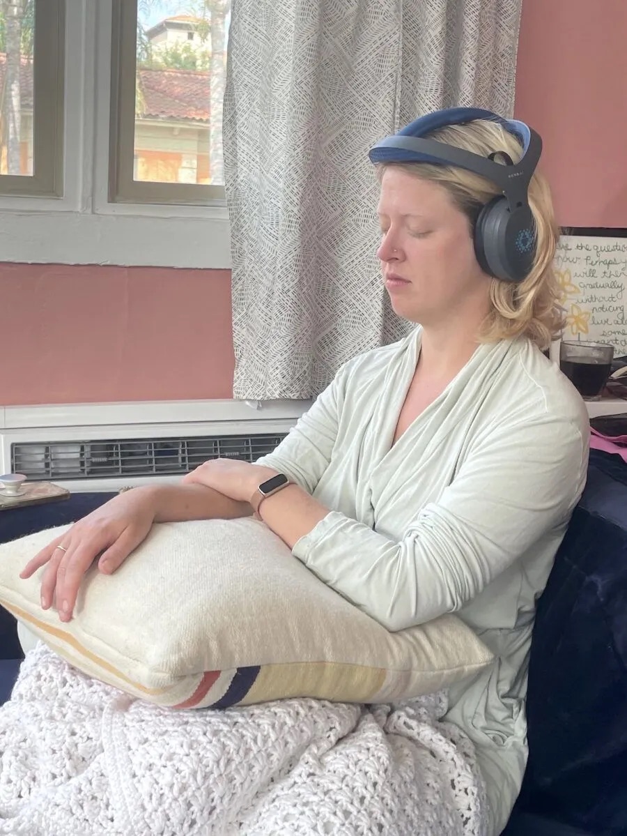 A woman sits on a couch with her eyes closed, wearing a headset device on her head and resting her arms on a pillow in her lap.