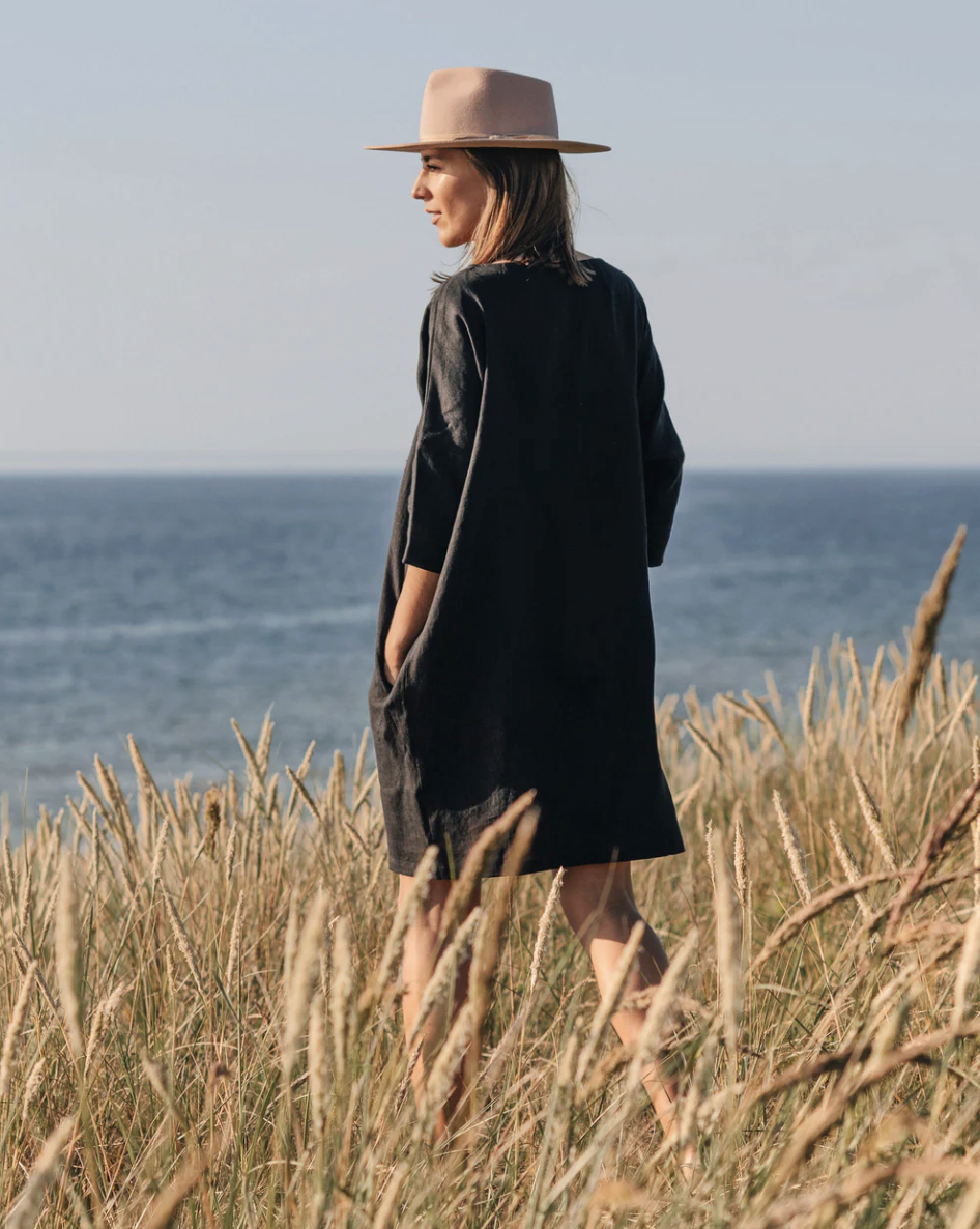 A person in a black dress and beige hat stands in tall grass, facing the sea under a clear sky.