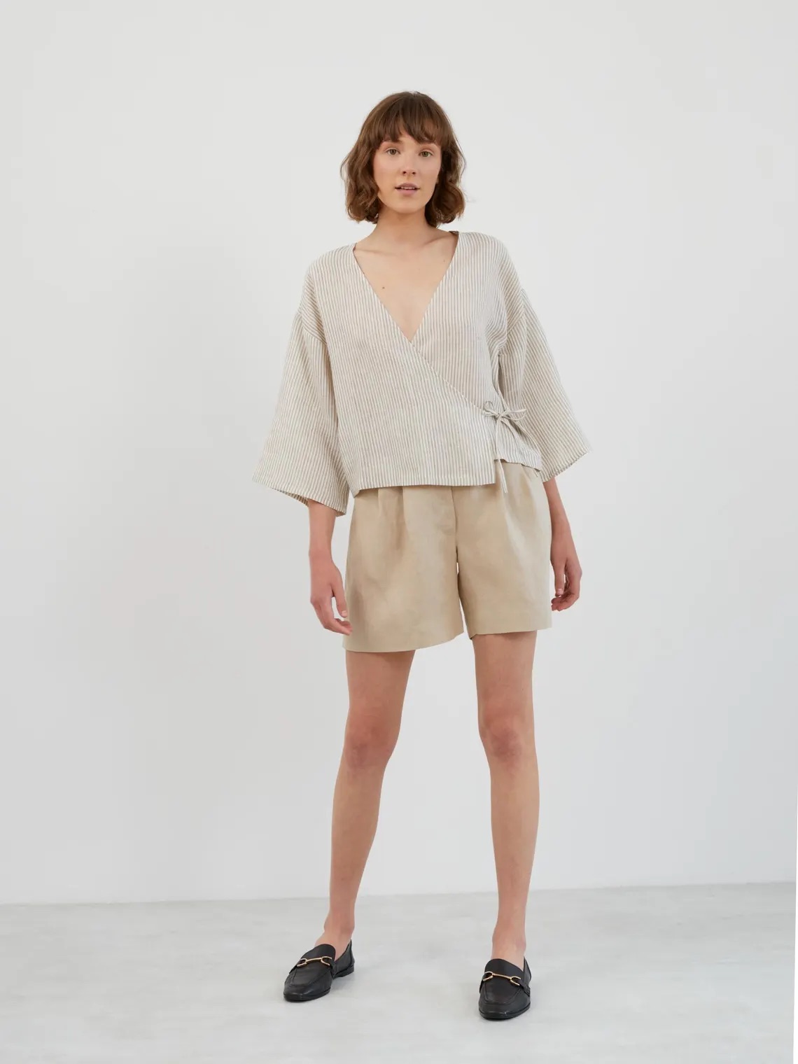 A woman stands against a plain white background wearing a loose, striped V-neck top, beige shorts, and black slip-on shoes.