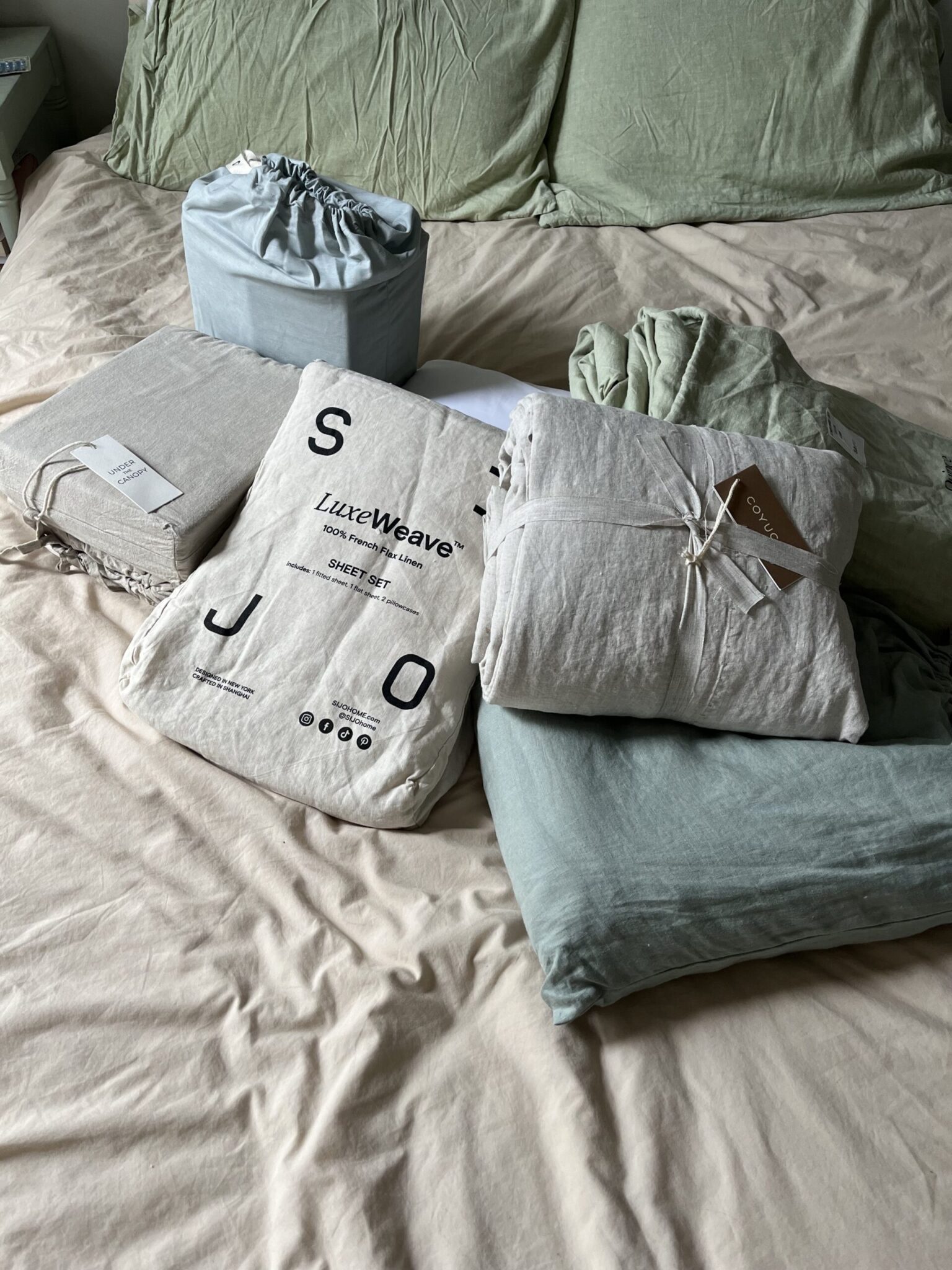 A variety of packaged bedding items, including sheet sets and pillowcases in green and beige tones, are arranged on a made bed with green pillows.
