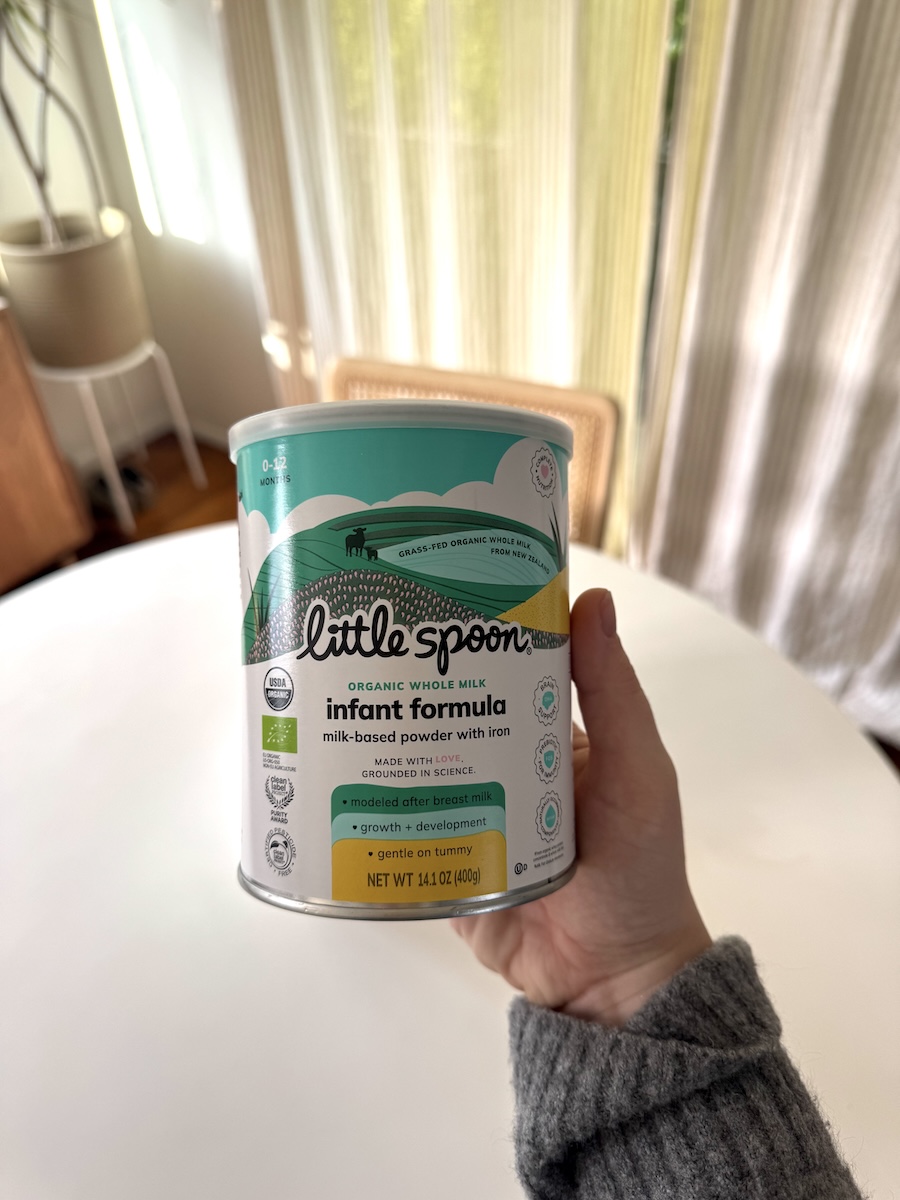 A hand holds a canister of Little Spoon organic whole milk infant formula in front of a white table and curtains in a sunlit room.