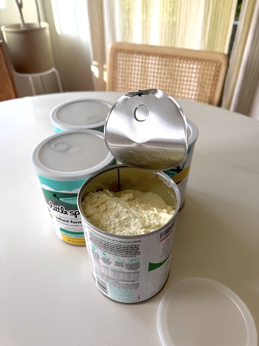 An open can of powdered infant formula sits on a table next to two unopened cans and a removed plastic lid, with a chair and window in the background.