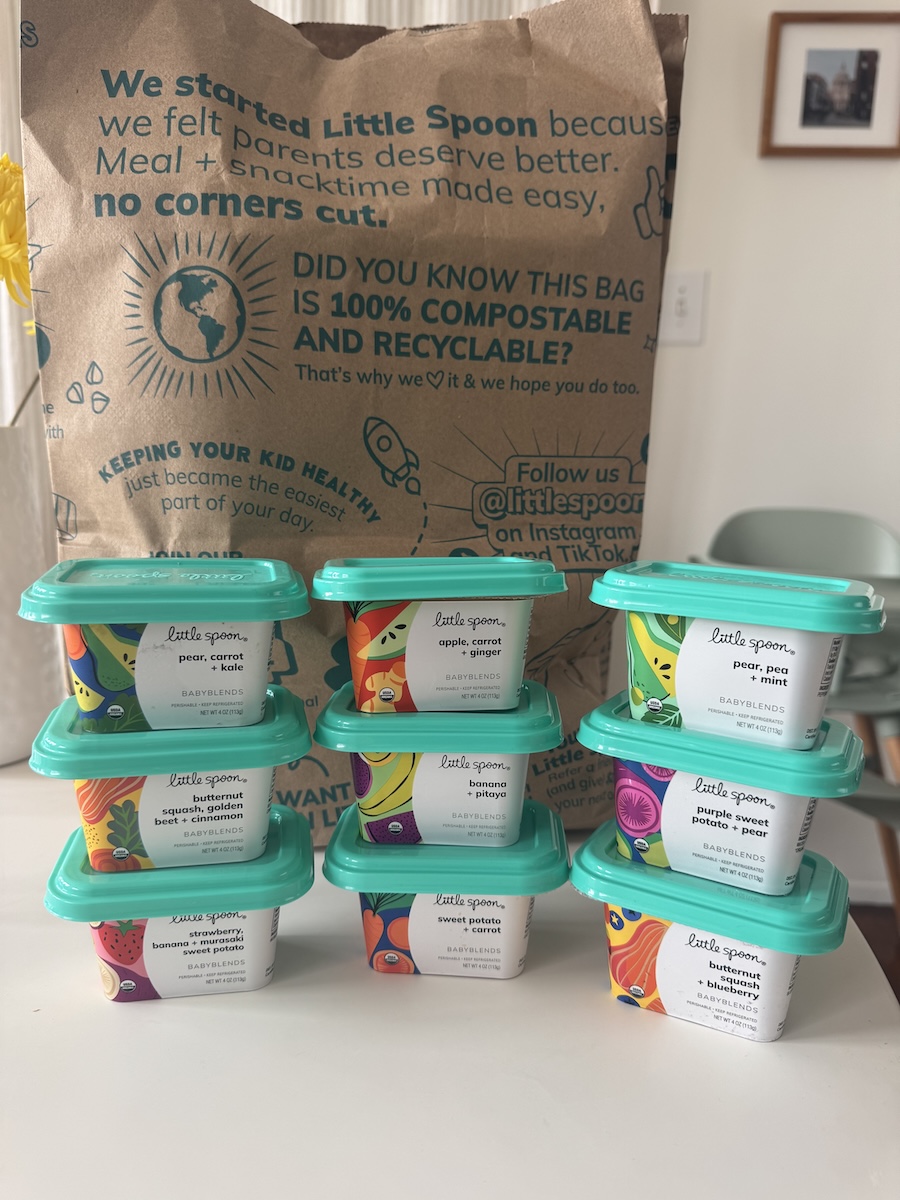 Eight containers of Little Spoon baby food are stacked on a table in front of a brown paper bag featuring eco-friendly messaging and branding.