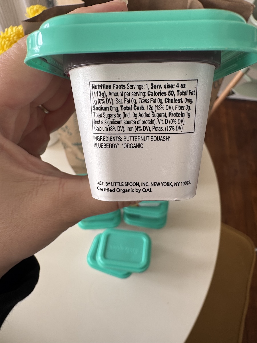 A hand holds a Little Spoon baby food container with a teal lid, showing the nutrition facts and ingredients label on the side.