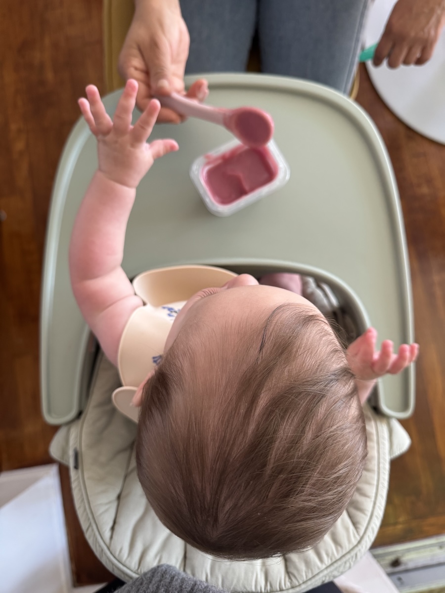 A baby in a high chair reaches toward a spoonful of pink baby food being offered by an adult.