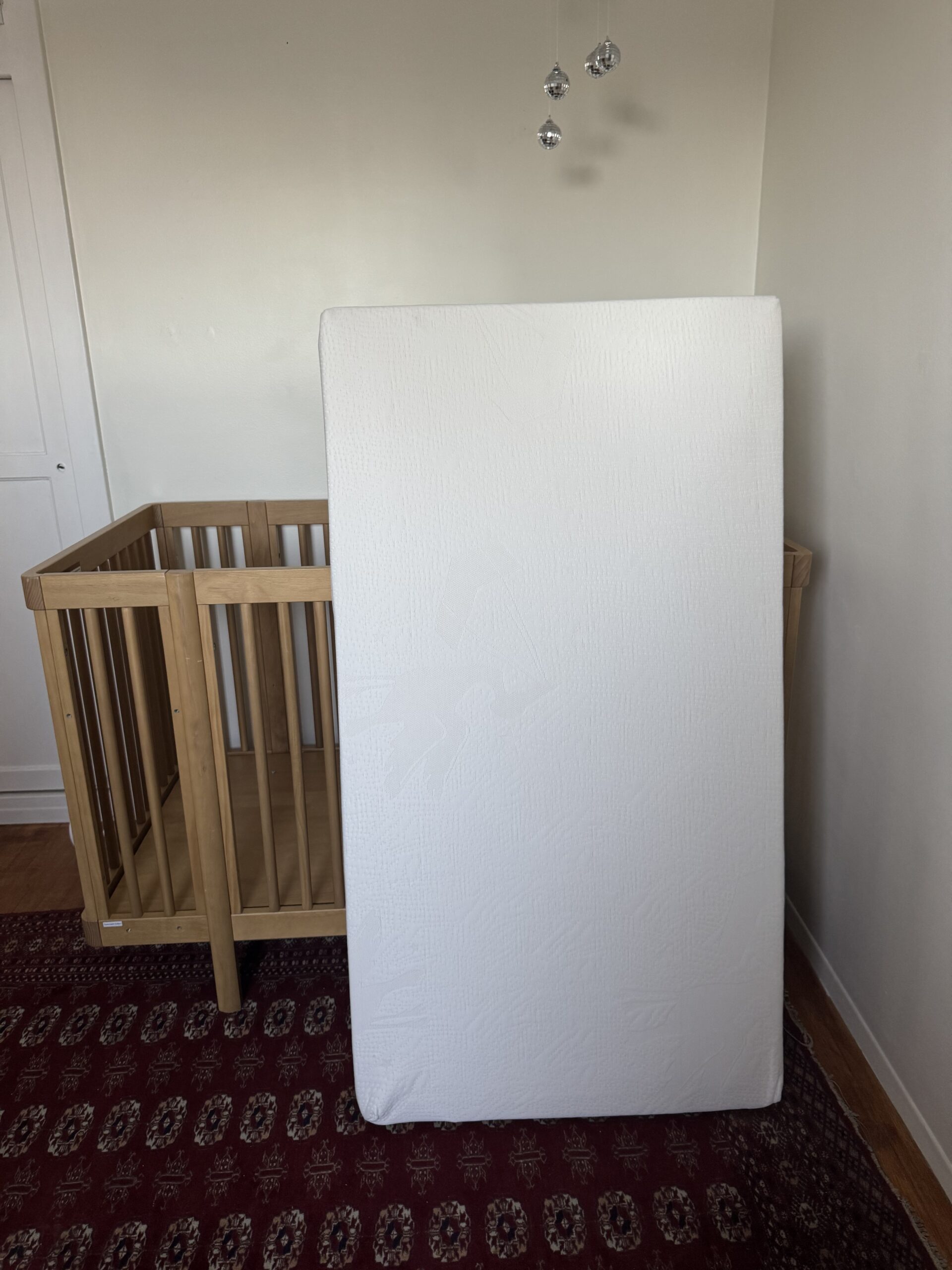 A crib mattress is propped upright against a wooden crib in a room with a patterned rug and light-colored walls.