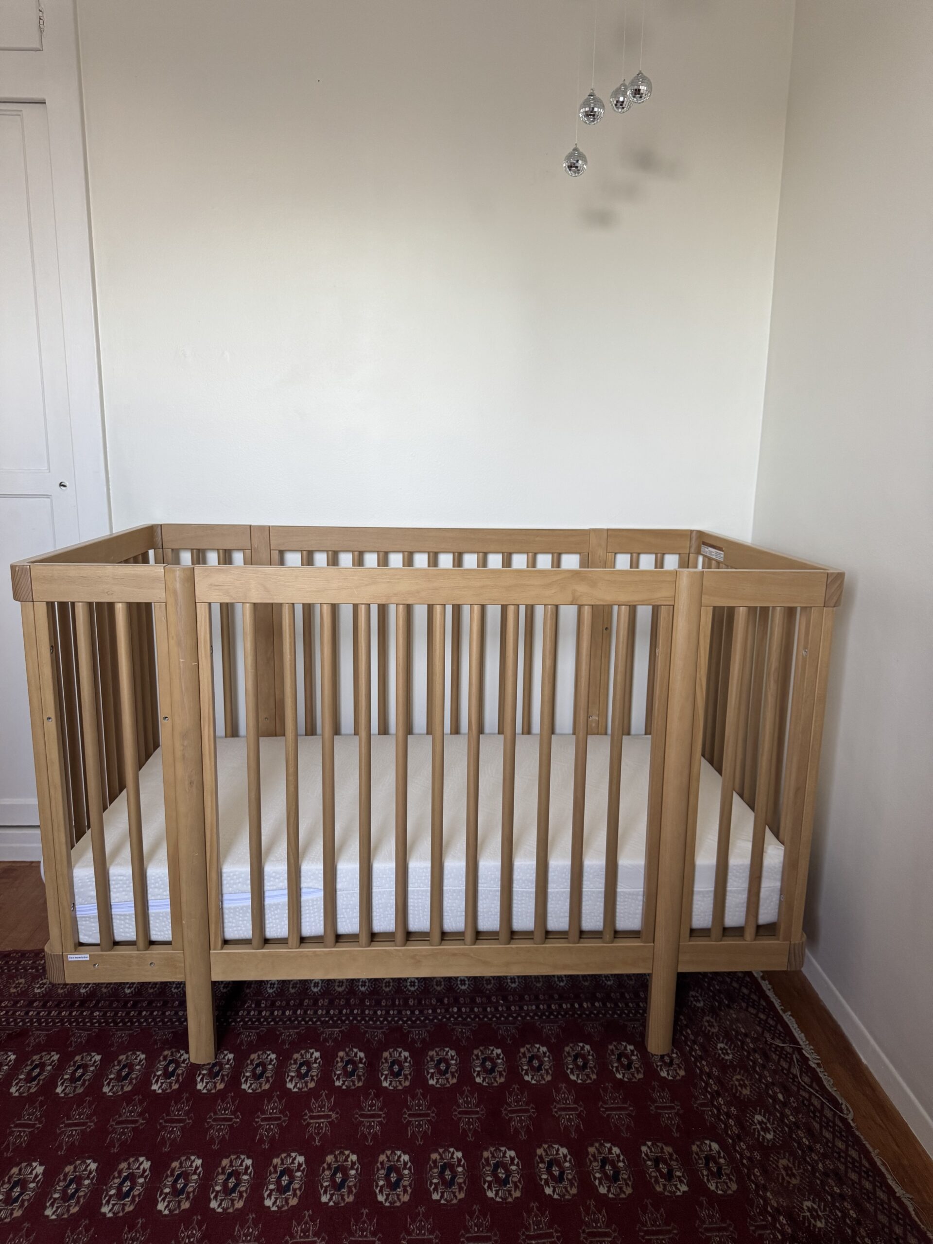 A wooden baby crib with a white mattress stands on a patterned rug against a plain wall, with a few small hanging decorations above.