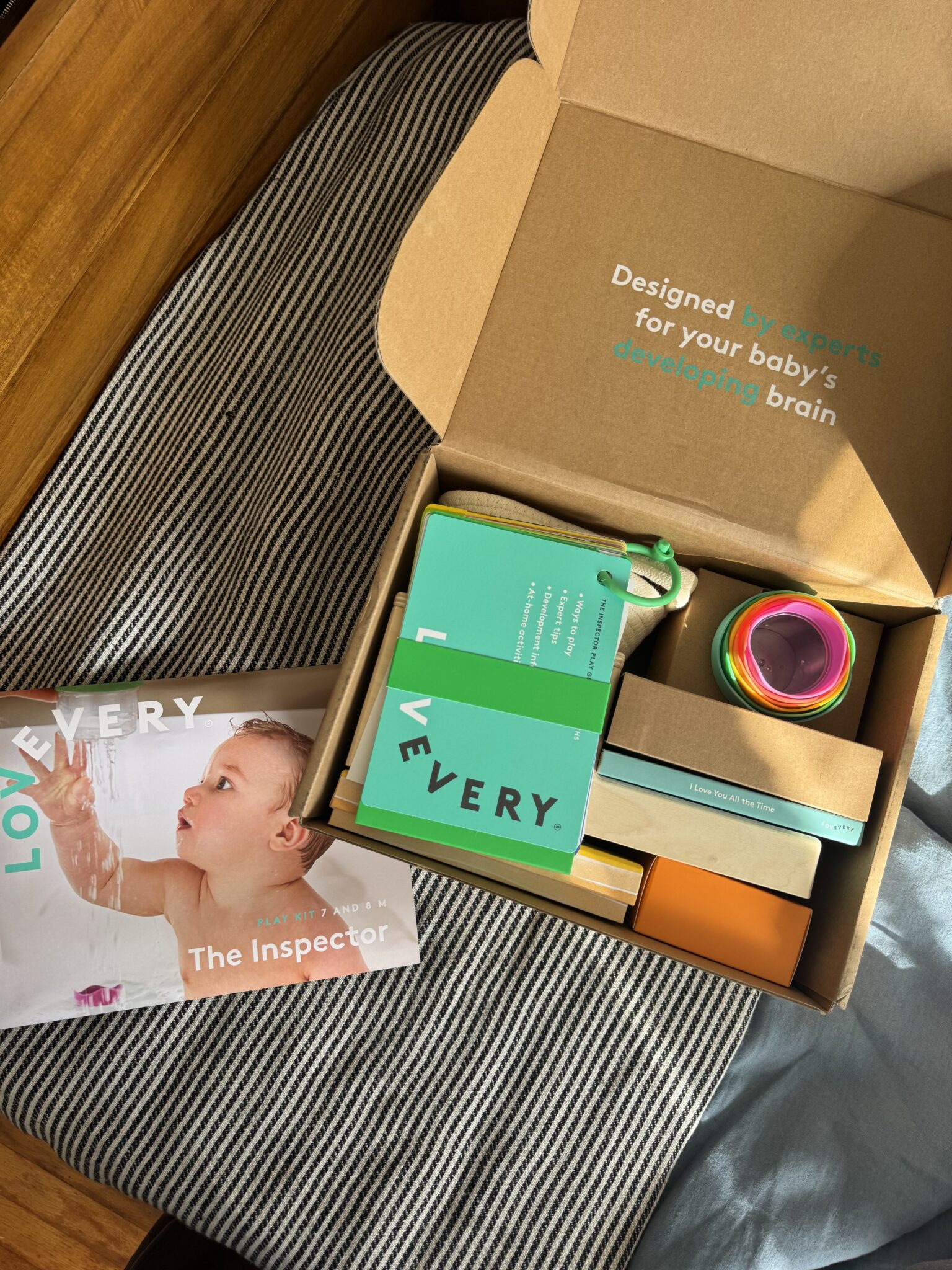 An open Lovevery play kit box with toys and materials inside, next to a booklet titled "The Inspector" featuring a baby on the cover, all placed on a striped cushion.