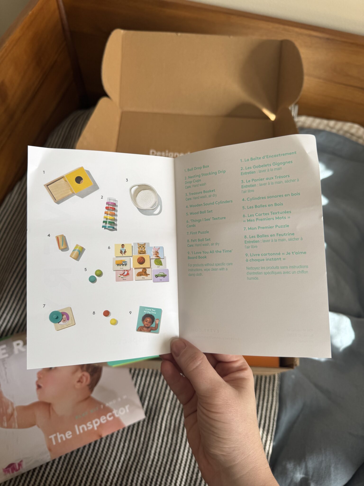 A hand holds open a booklet showing a list of items in French and images of small toys; a cardboard box and part of another booklet are visible in the background.