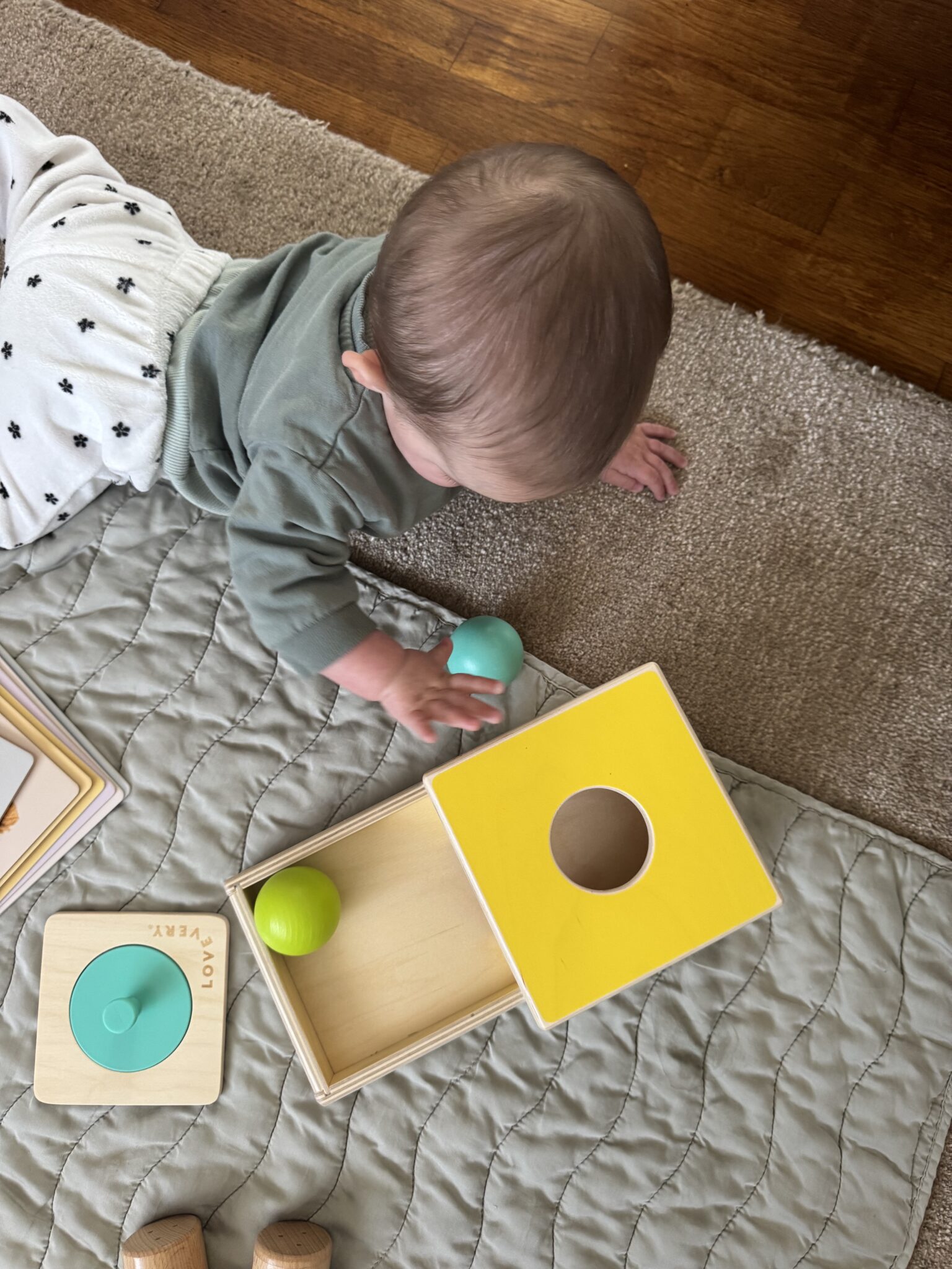 A baby is lying on a quilted mat, reaching for a green ball next to a wooden toy box with a yellow lid that has a circular hole.