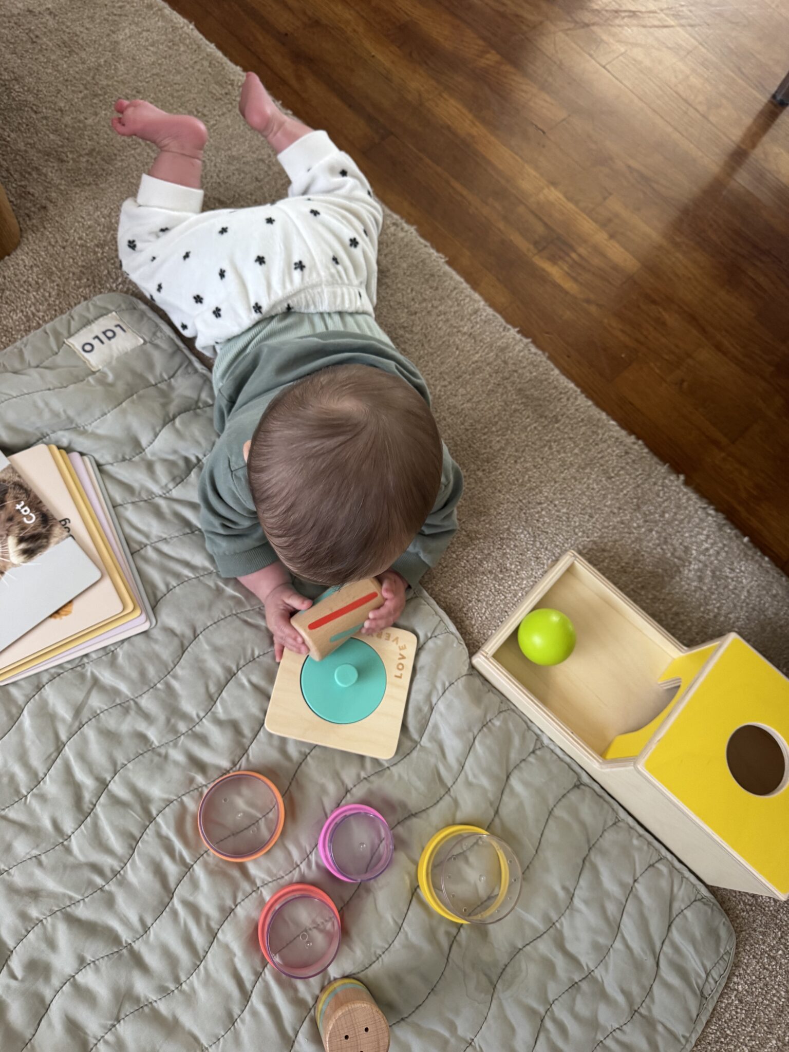 A baby lies on a quilted mat, playing with wooden stacking toys and colorful rings, next to a box with a green ball on a carpeted floor.