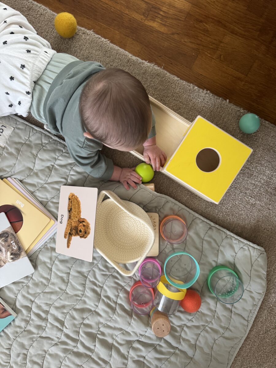 Baby lying on a mat playing with a yellow shape sorter, colorful cups, balls, and animal flashcards.