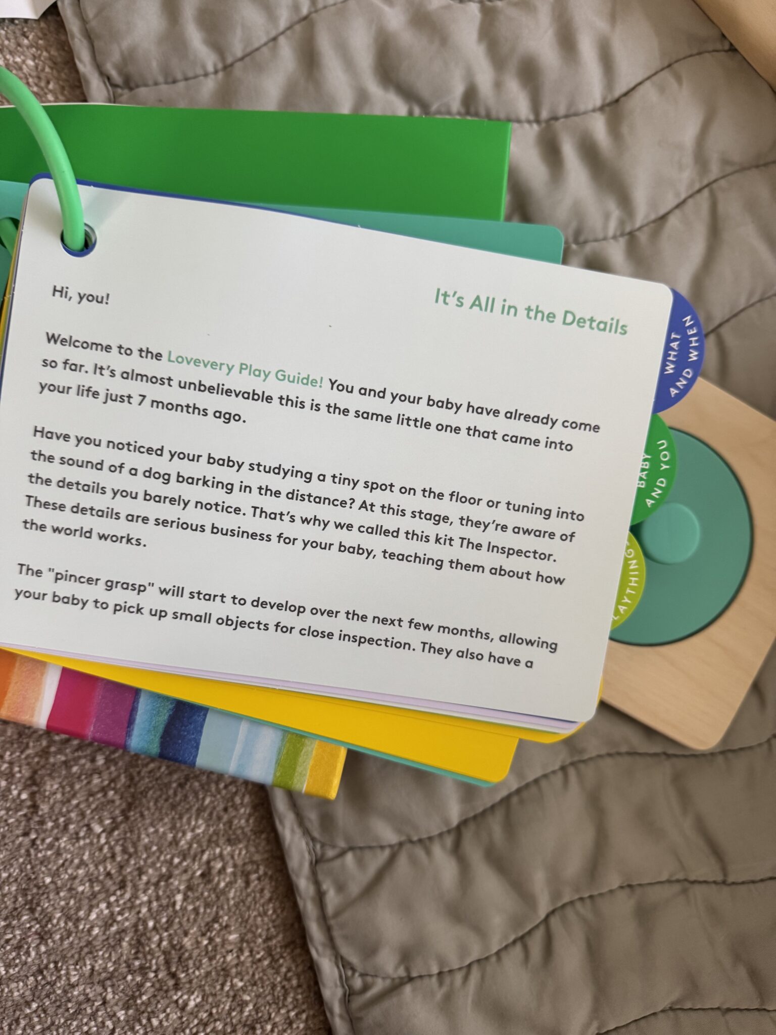 A card from the Lovevery Play Guide explains baby developmental milestones, with a focus on observation skills and the beginning of the pincer grasp. The card sits on a quilted blanket.