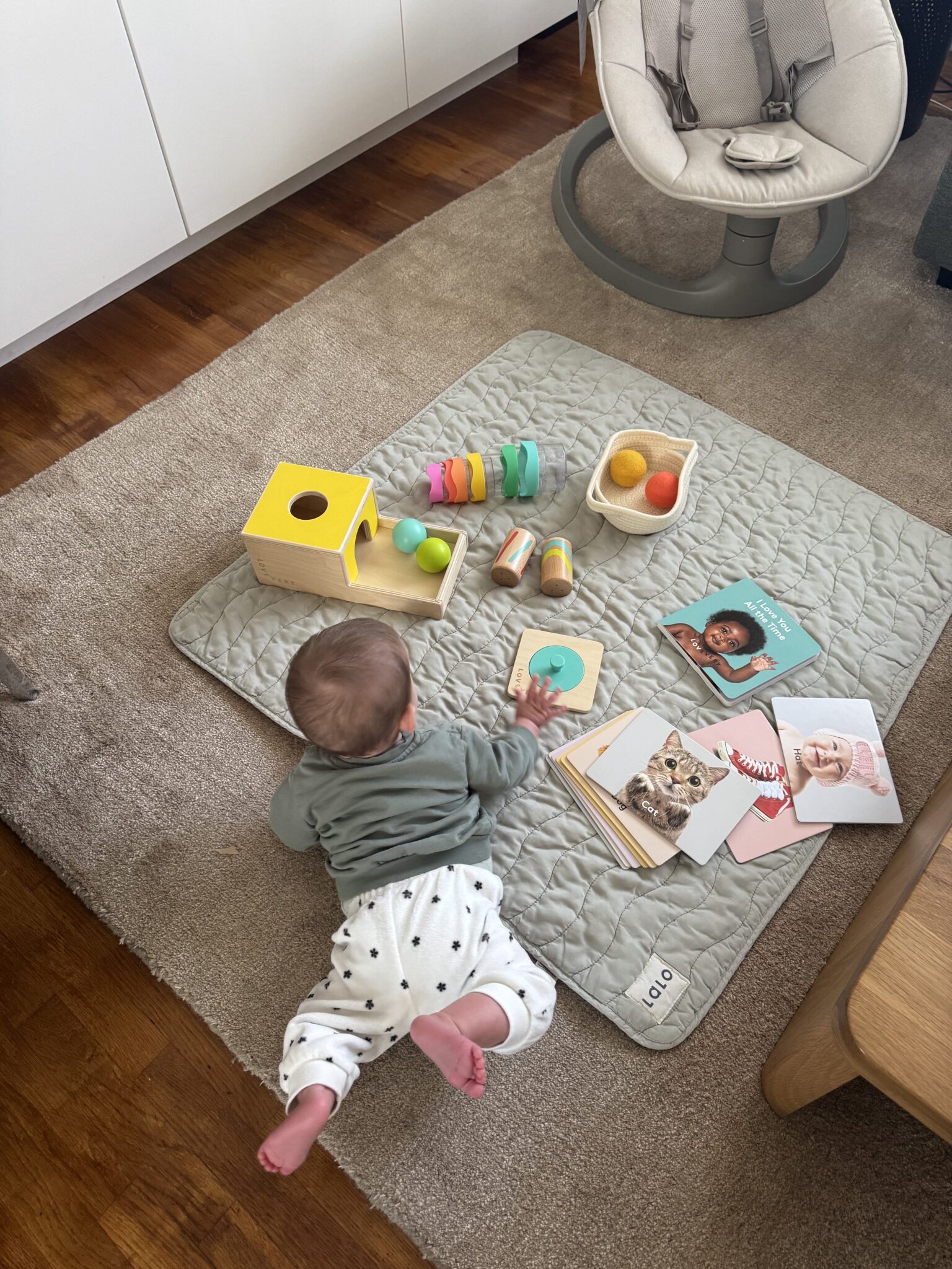 A baby lies on a quilted mat with wooden toys, colorful stacking rings, and animal and baby photo cards scattered around.