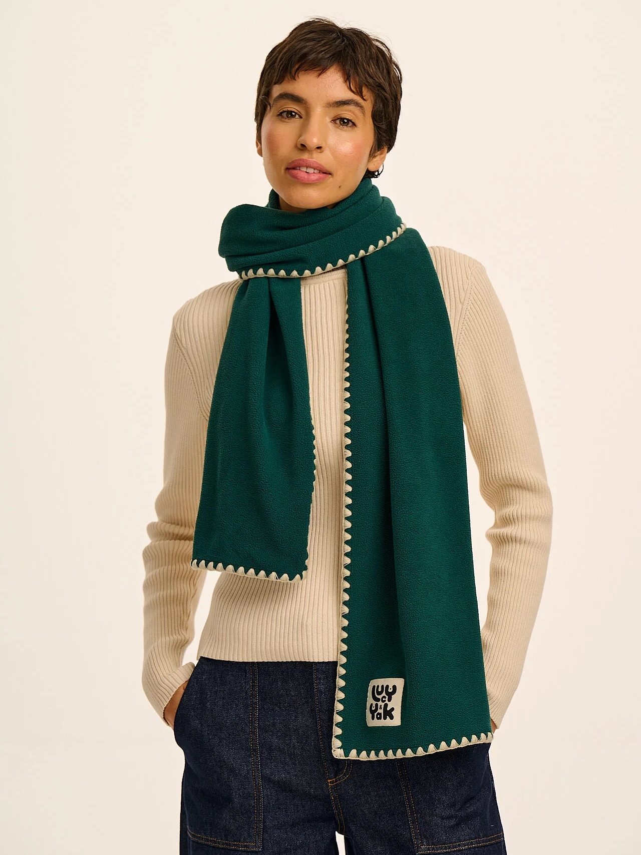 Person wearing a cream ribbed sweater, dark denim jeans, and a dark green scarf with white trim, standing against a plain light background.