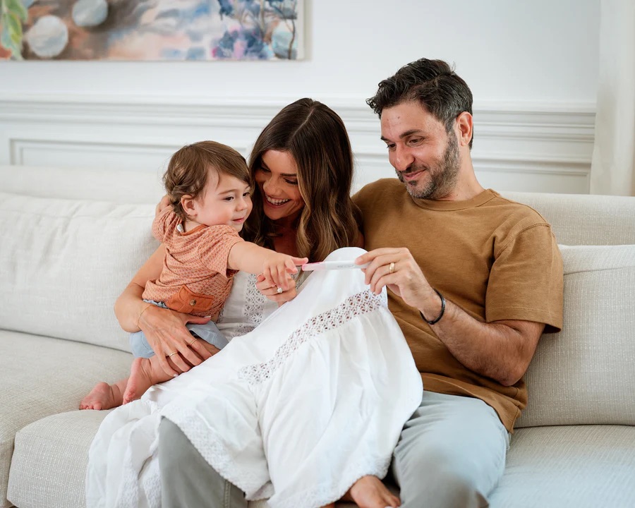 A man, woman, and young child sit on a couch together, smiling and holding a pregnancy test.