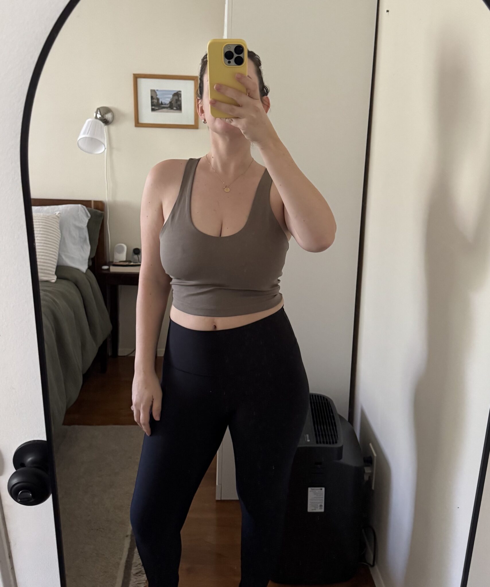 A person takes a mirror selfie in a bedroom, wearing a taupe crop top and black leggings, standing barefoot on a wooden floor.