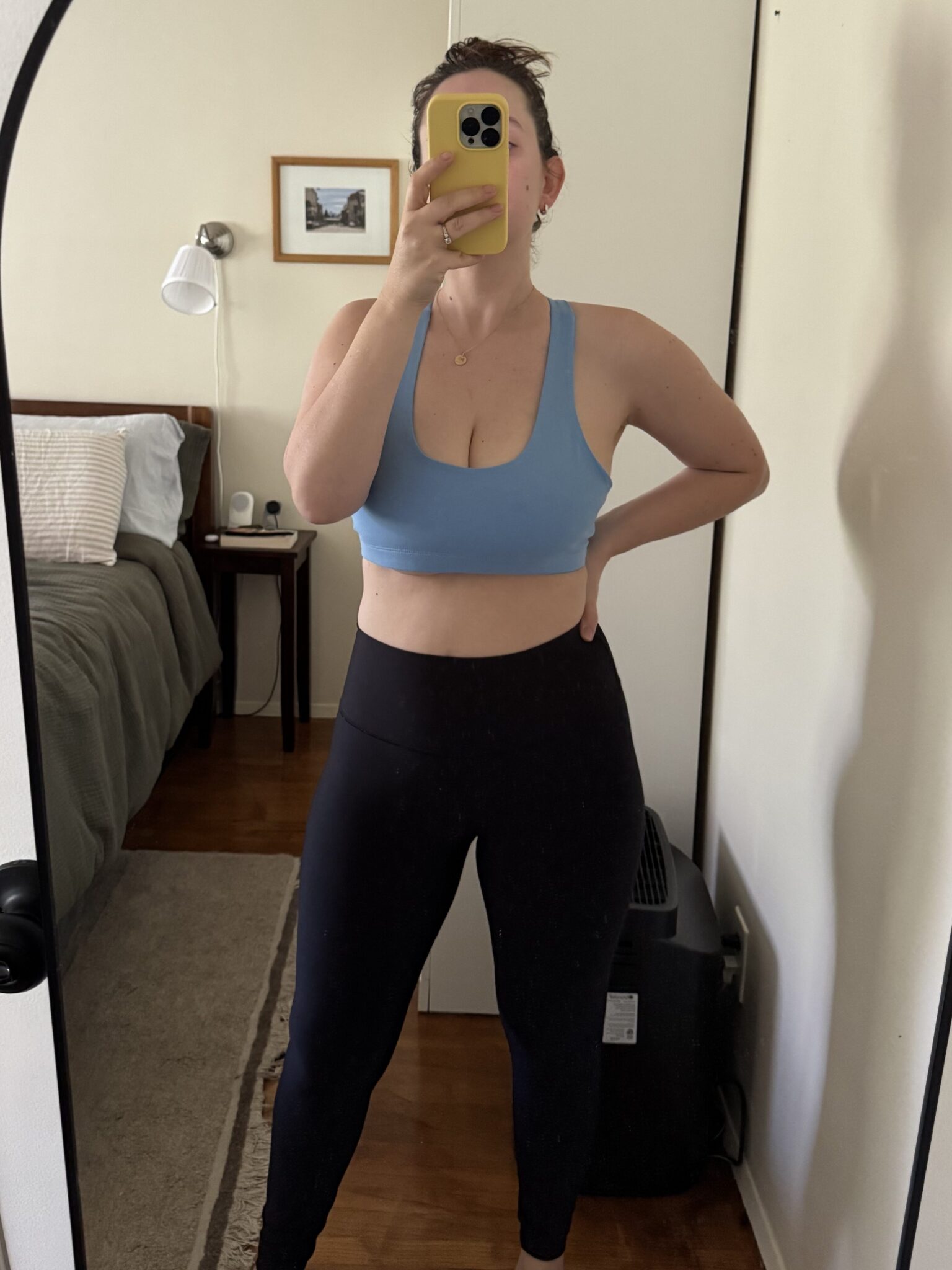 A person wearing a blue sports bra and black leggings takes a mirror selfie in a bedroom with a neatly made bed and wall art in the background.