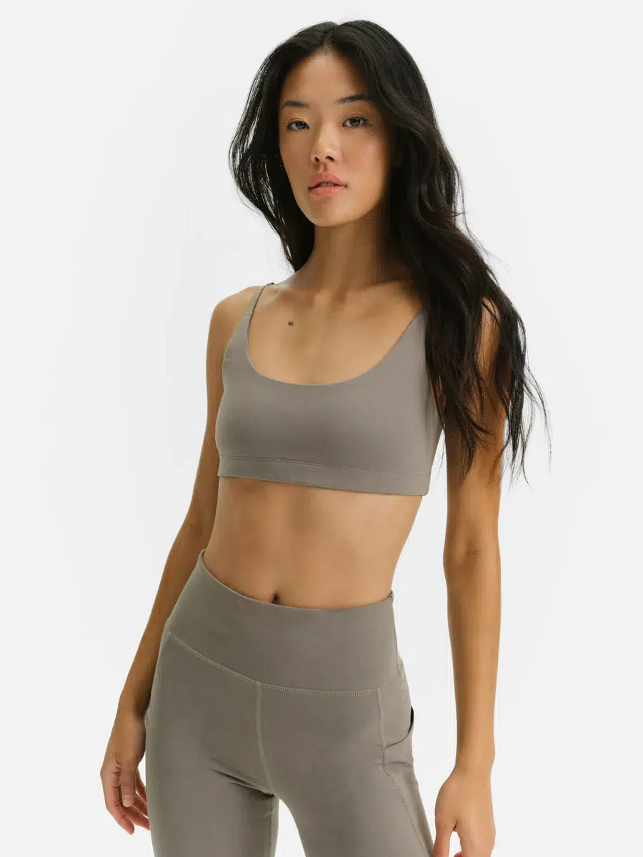 A woman stands against a plain background wearing a taupe sports bra and matching high-waisted leggings.