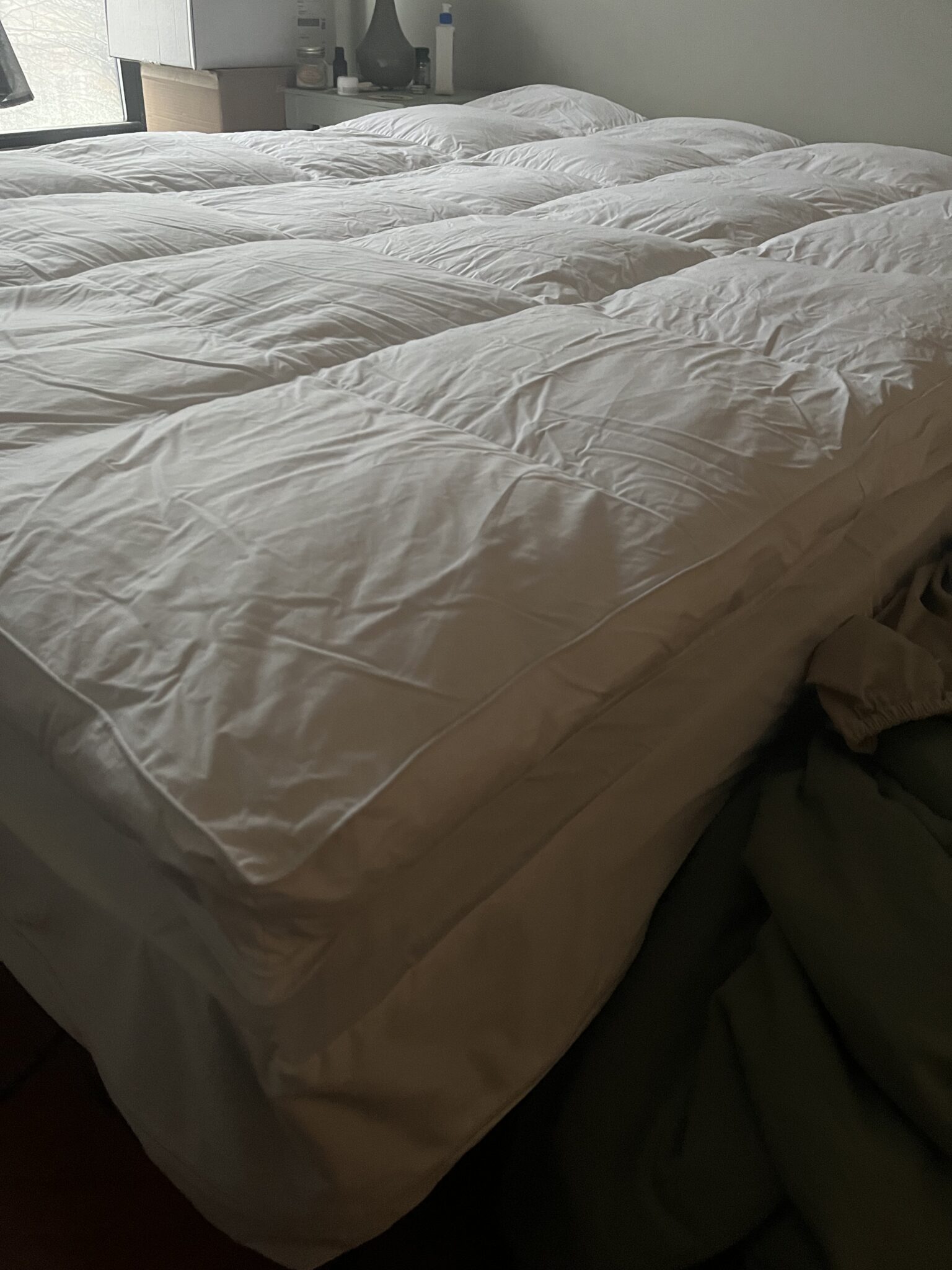 A neatly made bed with a white comforter in a dimly lit room, with a nightstand and a green blanket on the floor.