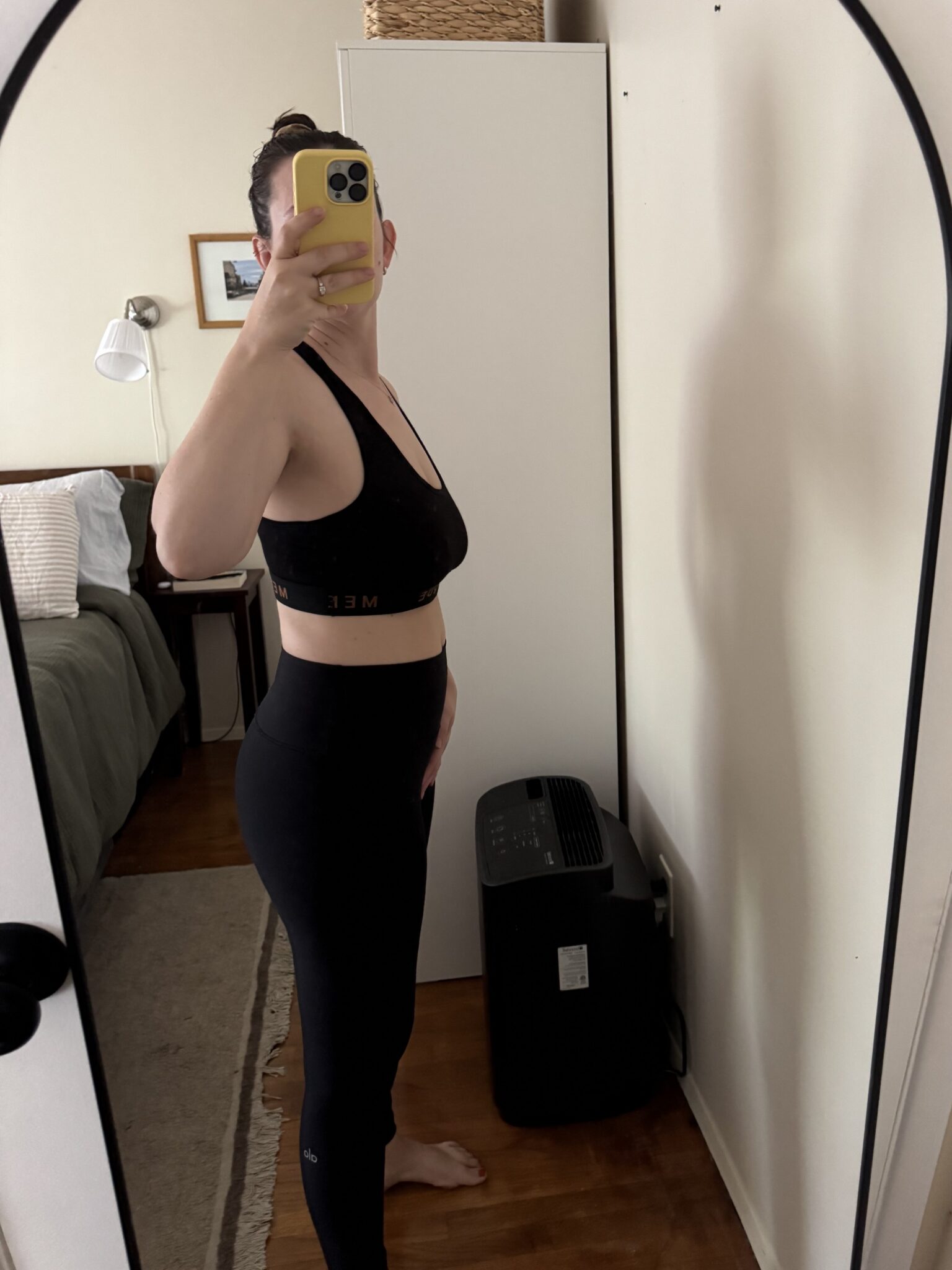 Person wearing black athletic wear stands sideways in front of a mirror, taking a photo with a yellow phone in a bedroom with a white cabinet and a bed visible.