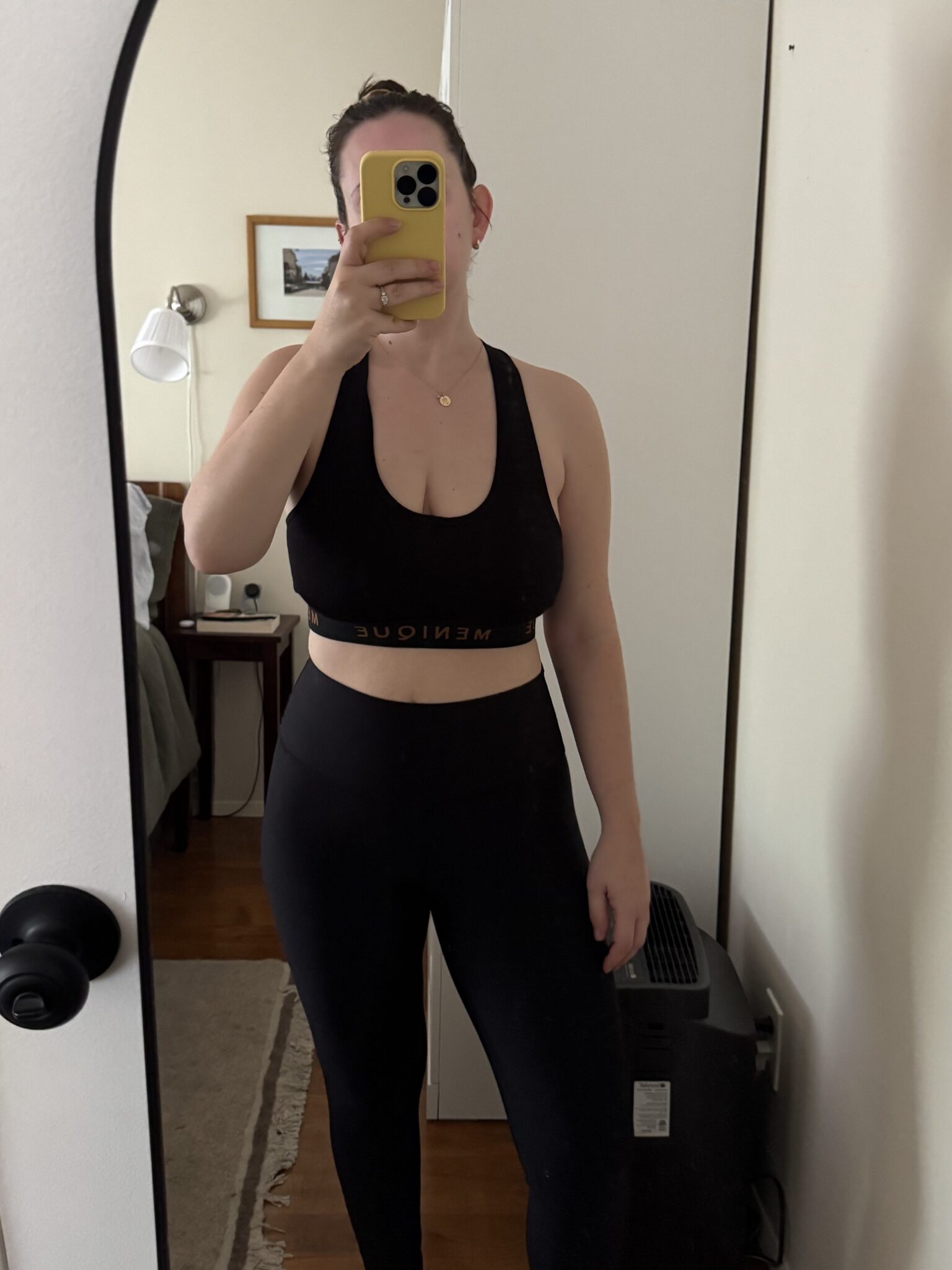 Person taking a mirror selfie in a room, wearing a black sports bra and leggings. A basket is on top of a cabinet, and part of a bed and nightstand are visible in the background.