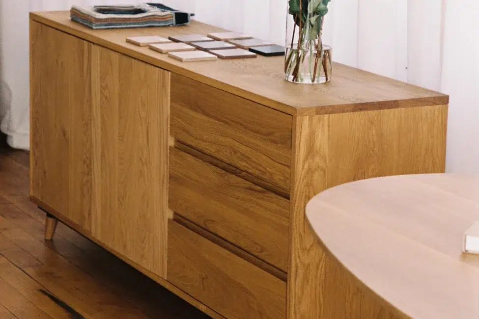 A wooden sideboard with two cabinet doors and three drawers, holding stacked books, sample tiles, and a glass vase with flowers, next to a round table on a hardwood floor.