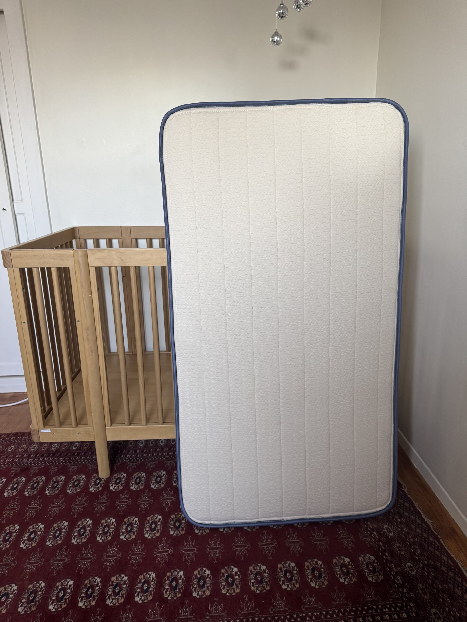 A wooden baby crib is next to a white crib mattress standing upright on a red patterned rug in a room with cream-colored walls.