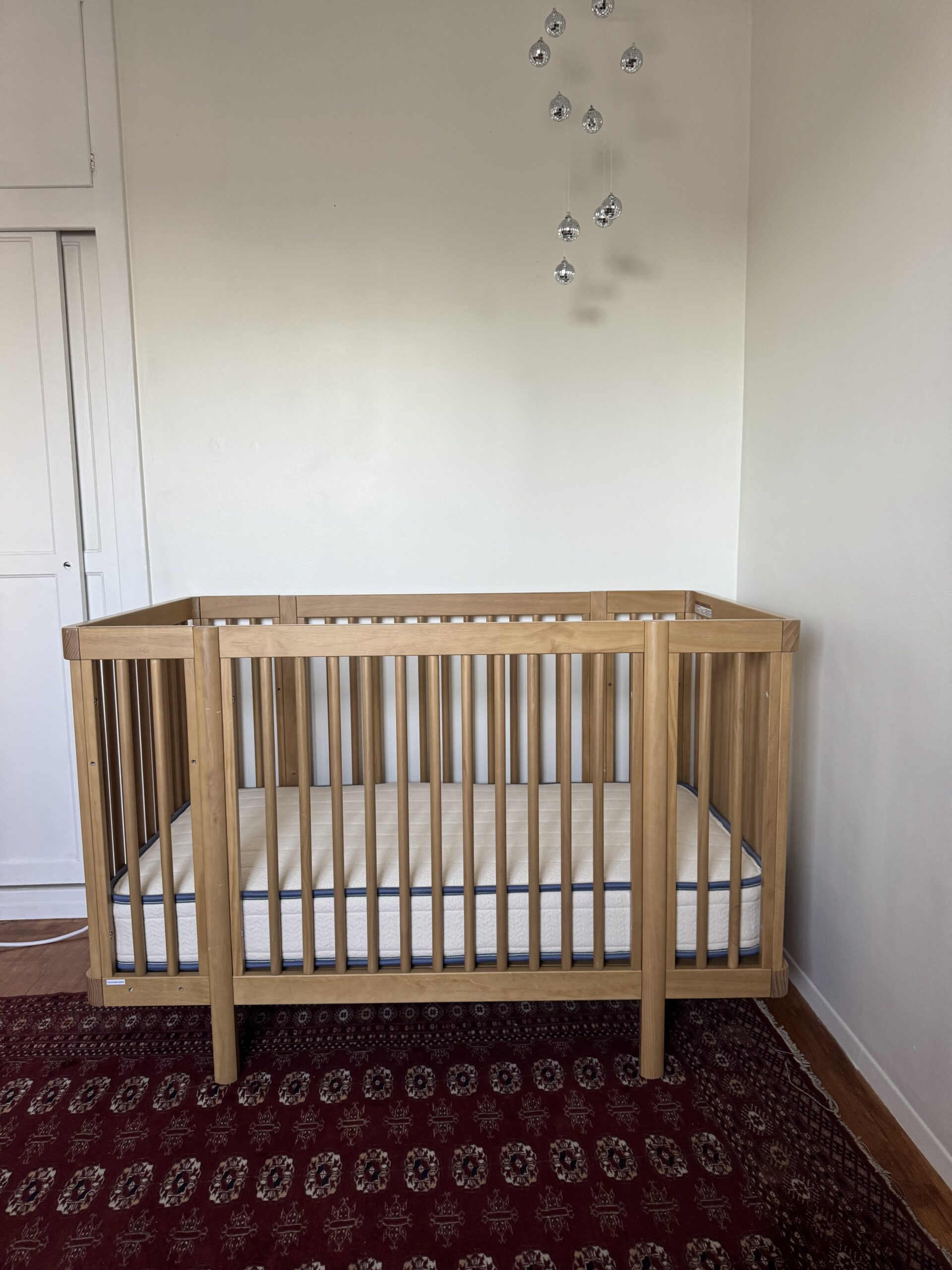 A wooden crib with a mattress is placed on a patterned rug against a white wall, with a small mobile of hanging crystals above it.