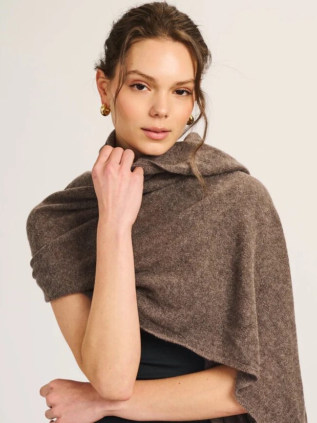 A woman wearing gold hoop earrings and a brown shawl draped over her shoulders stands against a plain background, looking at the camera.