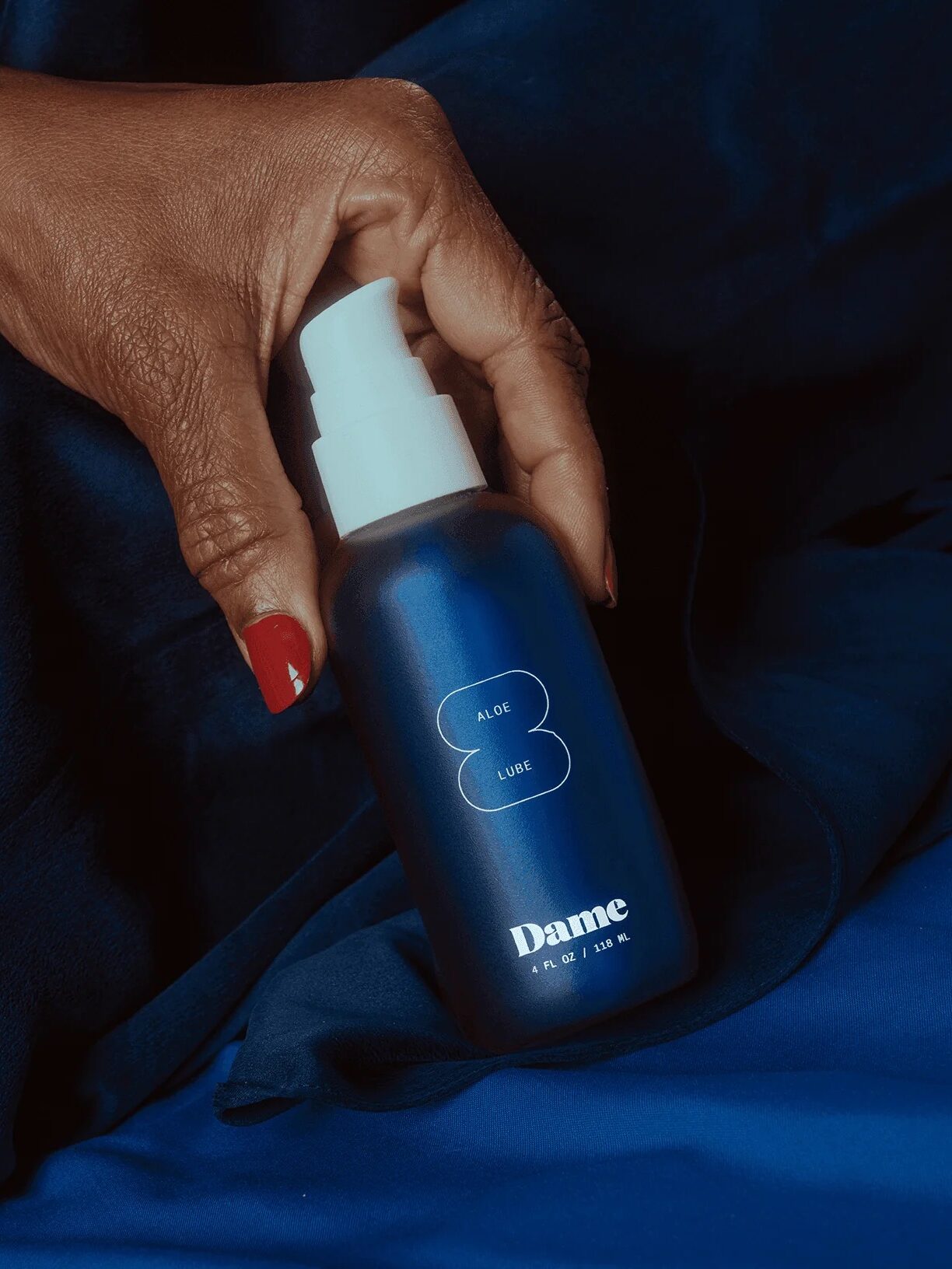 A hand with red nail polish holds a blue bottle labeled "Dame Aloe Lube" against a dark blue fabric background.