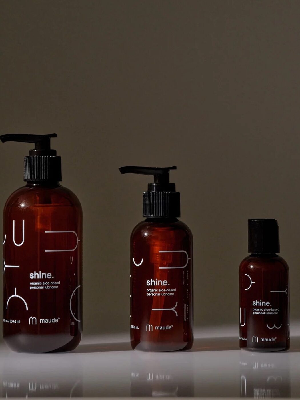 Three brown bottles of Maude's shine organic oil-based lubricant are arranged in a row against a plain background, with varying sizes from left to right.
