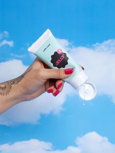 A hand with red nails and a tattoo holds an open tube of clear jelly against a blue sky with clouds.