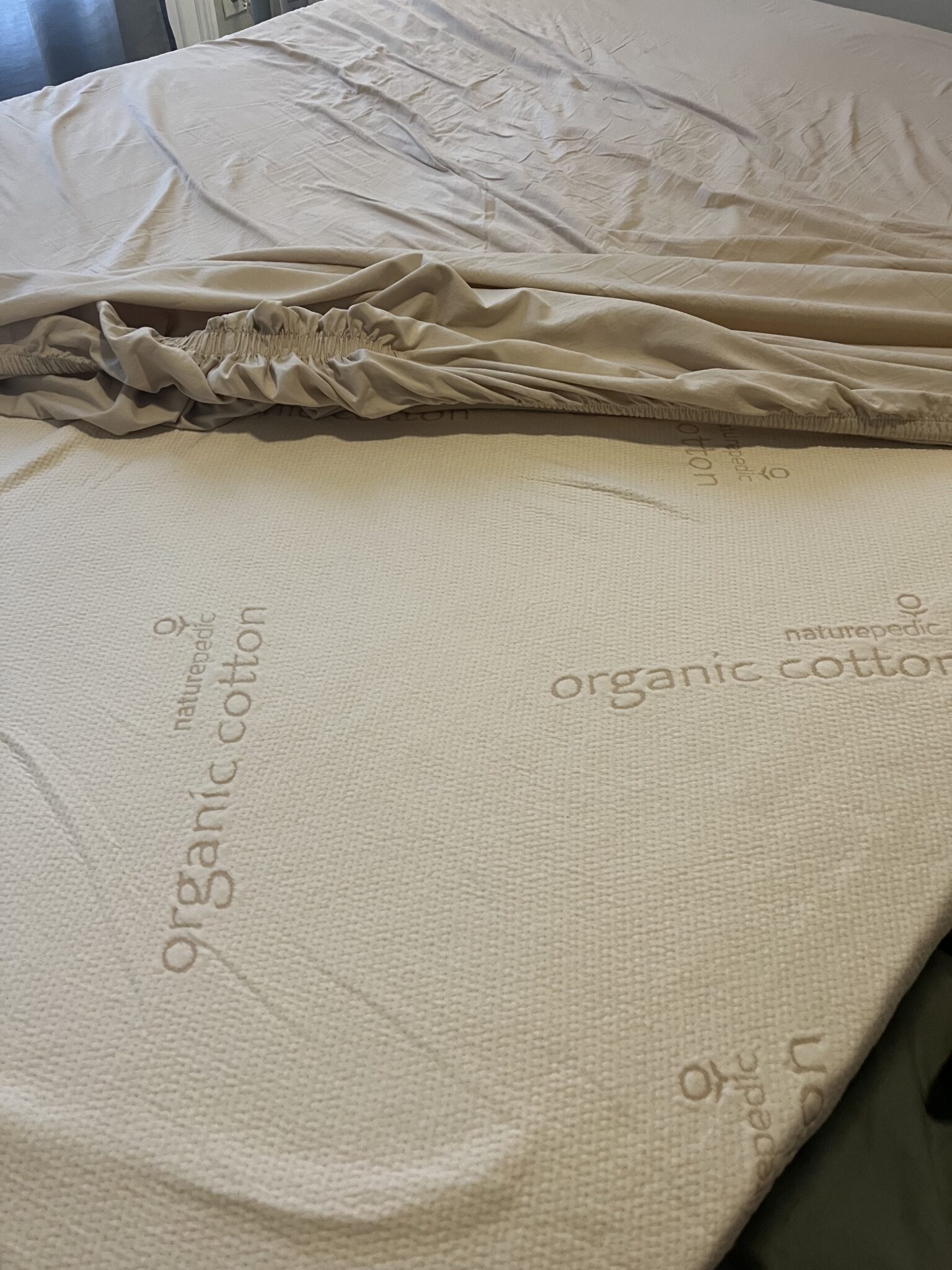 A beige fitted sheet is partially removed, exposing a mattress cover labeled "organic cotton" and "naturepedic.