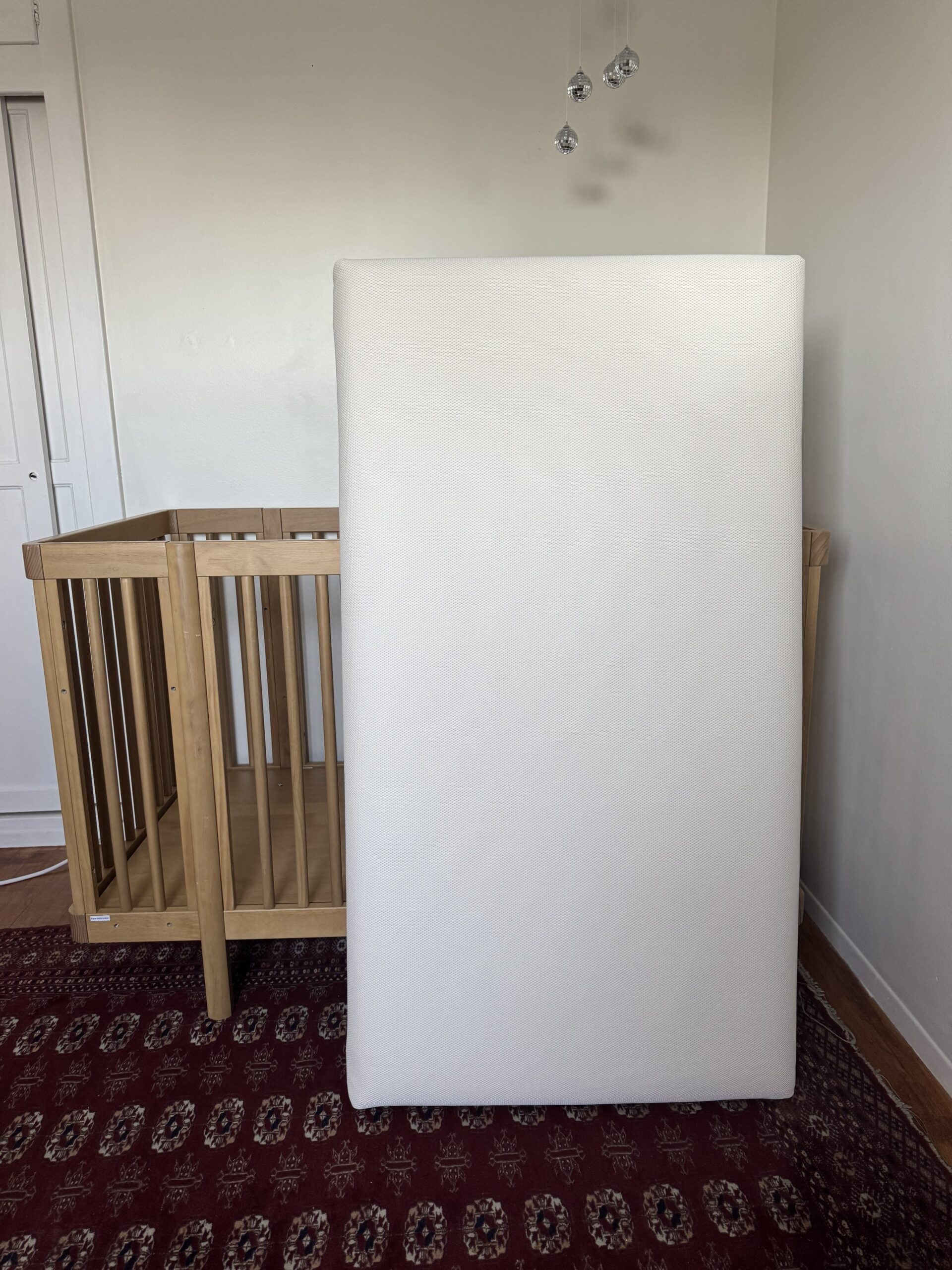 A crib mattress stands upright in front of an empty wooden crib on a red patterned rug in a minimally decorated room.