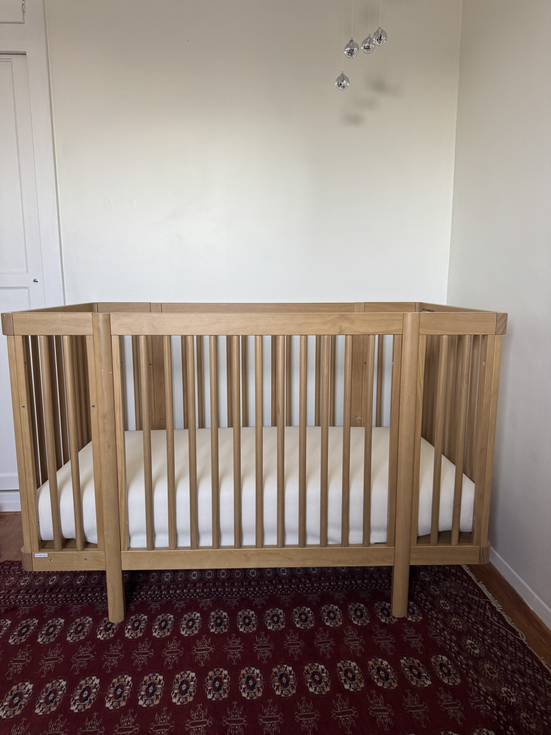 A wooden baby crib with a white mattress stands on a red patterned rug in a room with white walls and a closed white door.