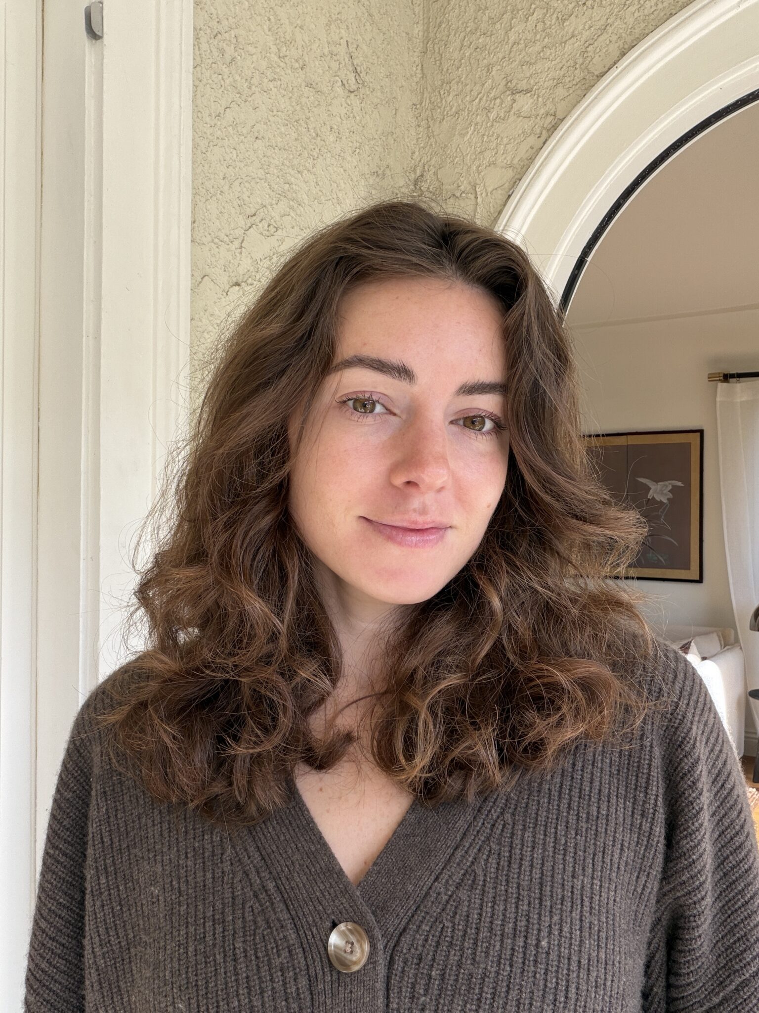 A woman with wavy brown hair and a brown sweater stands indoors near a white doorway, looking at the camera with a neutral expression.