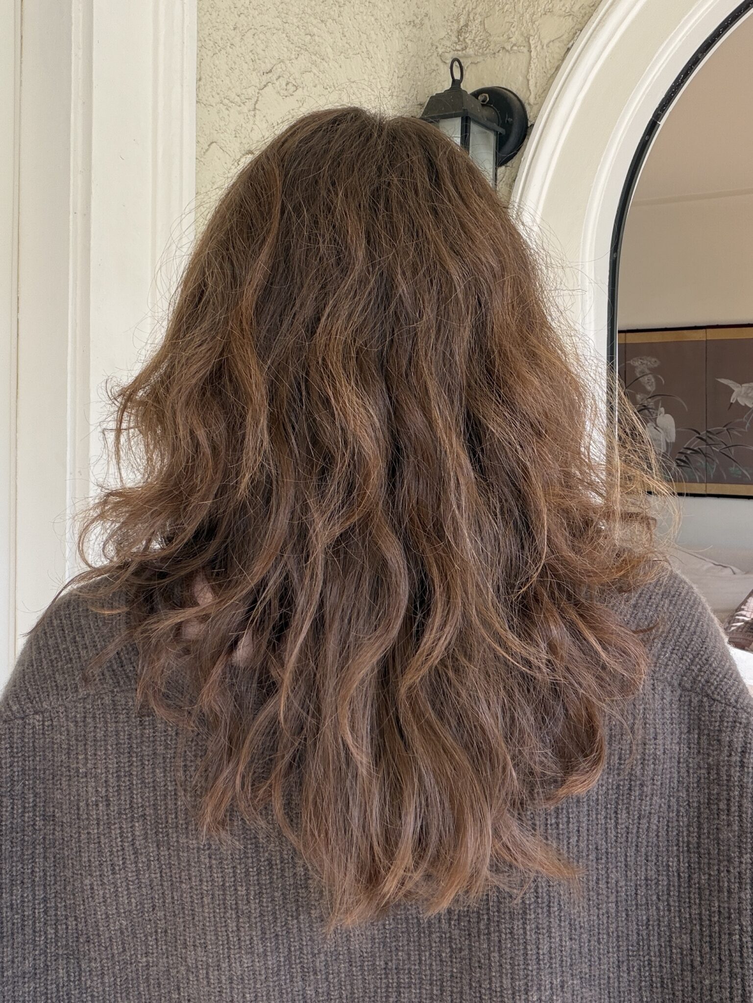 Person with medium-length, wavy brown hair seen from behind, wearing a brown sweater, standing indoors near a white doorway and a mirror.