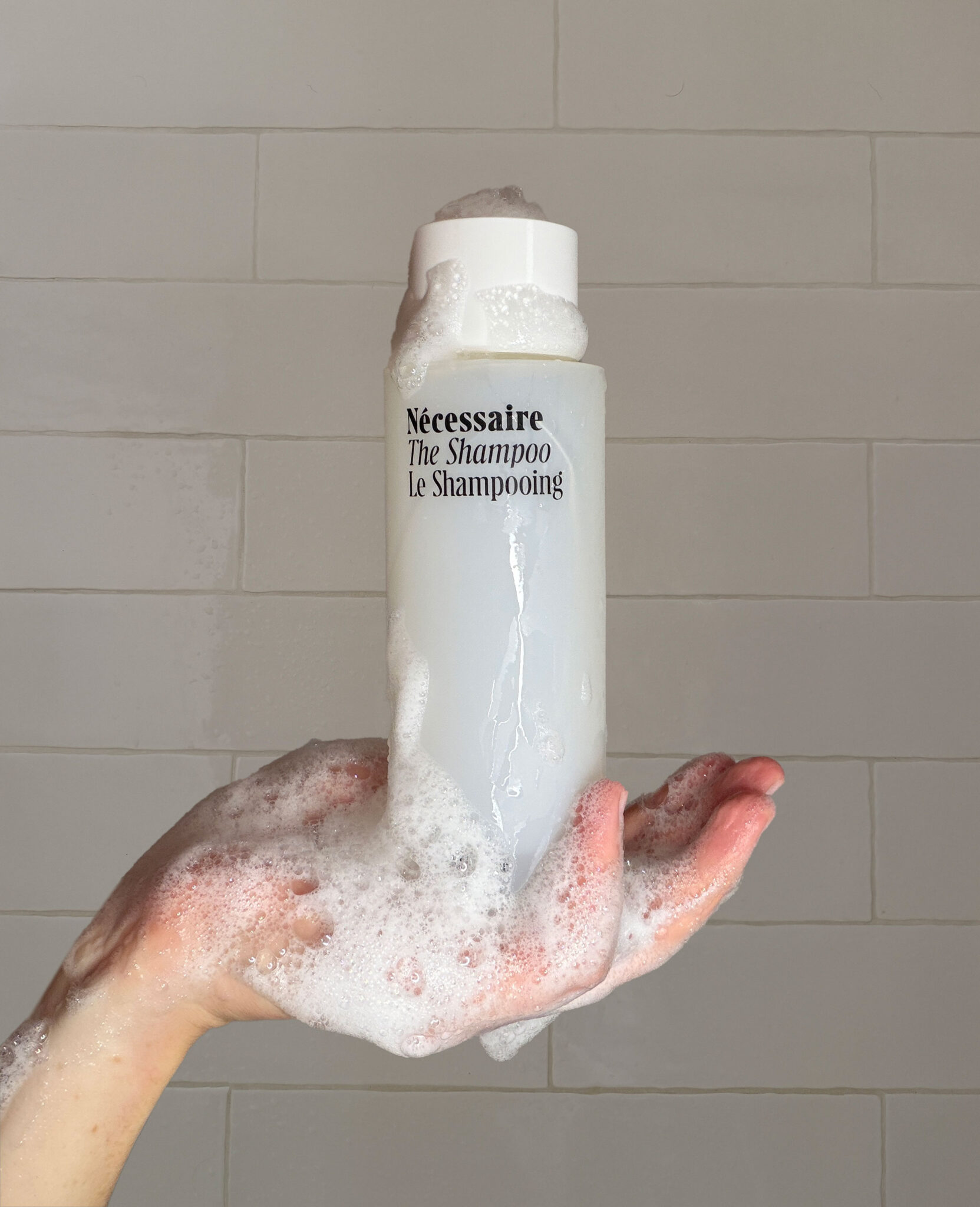 A hand covered in shampoo lather holds a bottle labeled “Nécessaire The Shampoo Le Shampooing” against a tiled background.