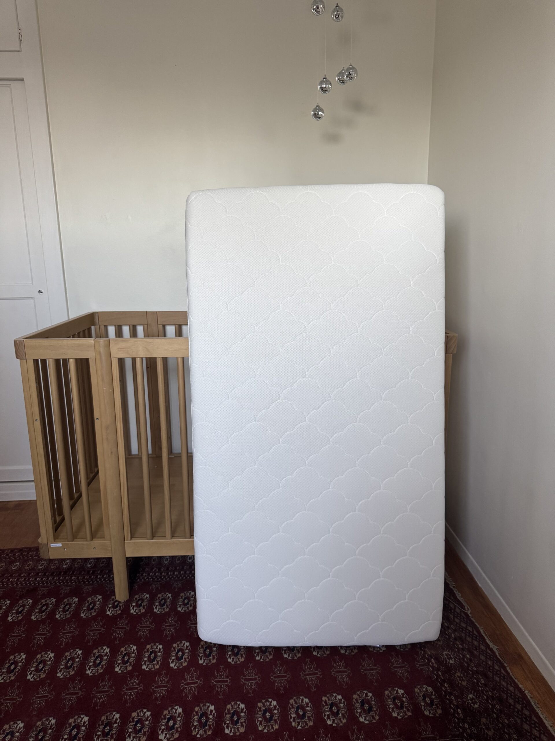 A white crib mattress stands upright in front of a wooden crib in a room with white walls and a patterned red rug.